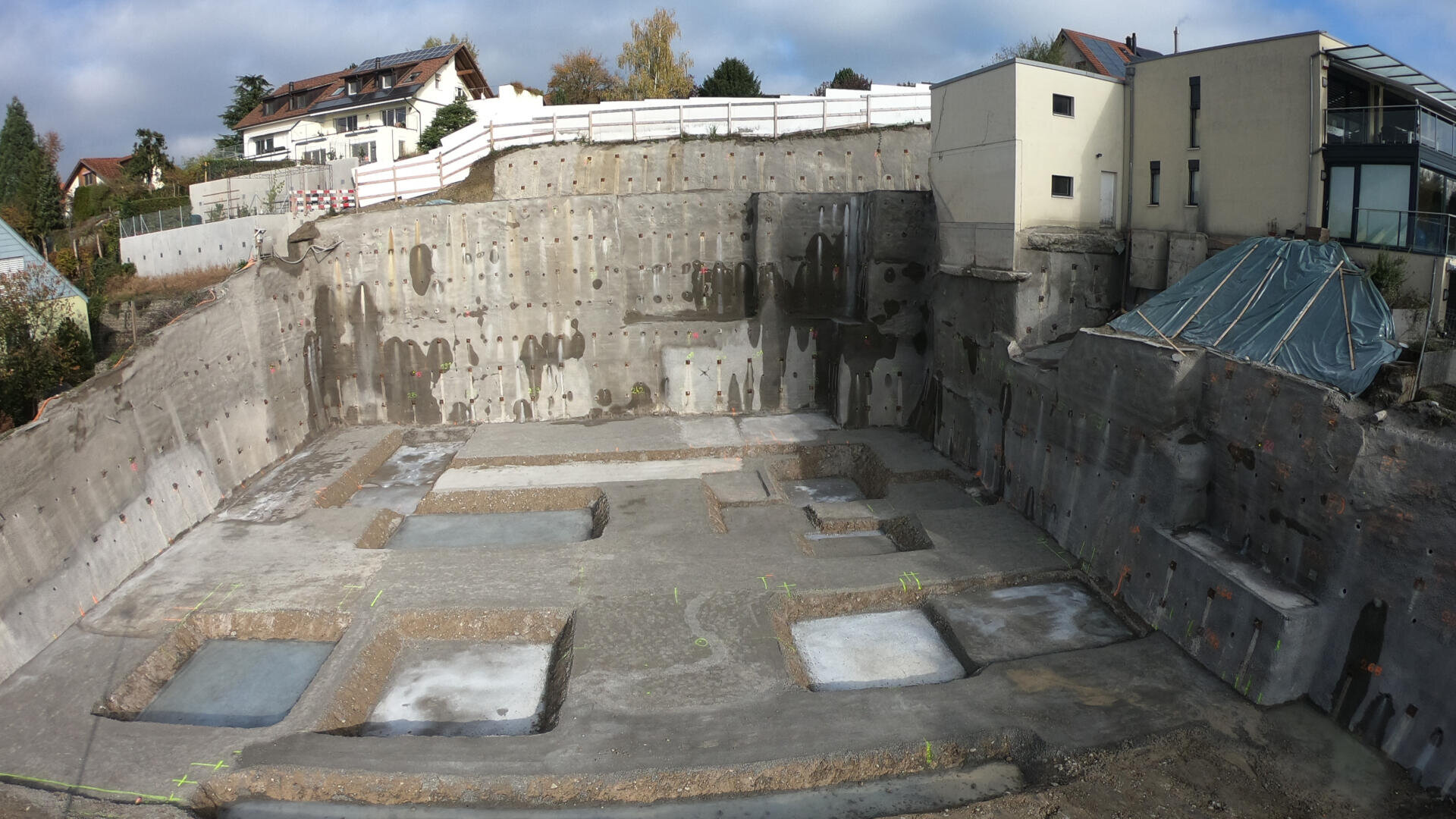 Eine Baustelle mit einer tiefen Baugrube, die von Betonstützmauern umgeben ist, mit auf dem Boden markierten Baugruben und im Hintergrund sichtbaren Wohngebäuden bei teilweise bewölktem Himmel.