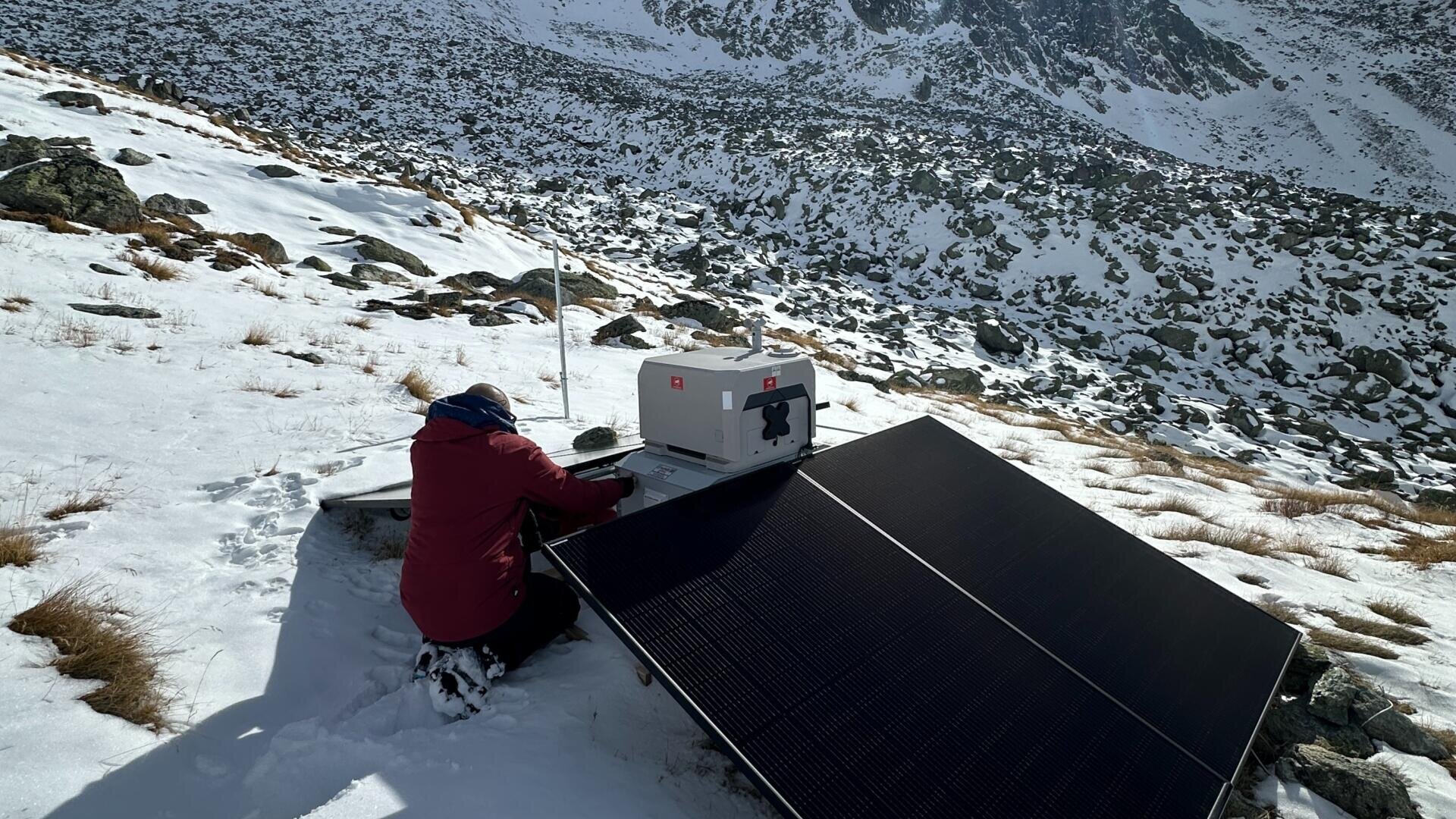 Eine Person in roter Jacke kniet auf dem verschneiten Boden und arbeitet an einem Solarmodul und einem Gerätekasten, die an einem Berghang mit felsigem, schneebedecktem Gelände im Hintergrund aufgestellt sind.