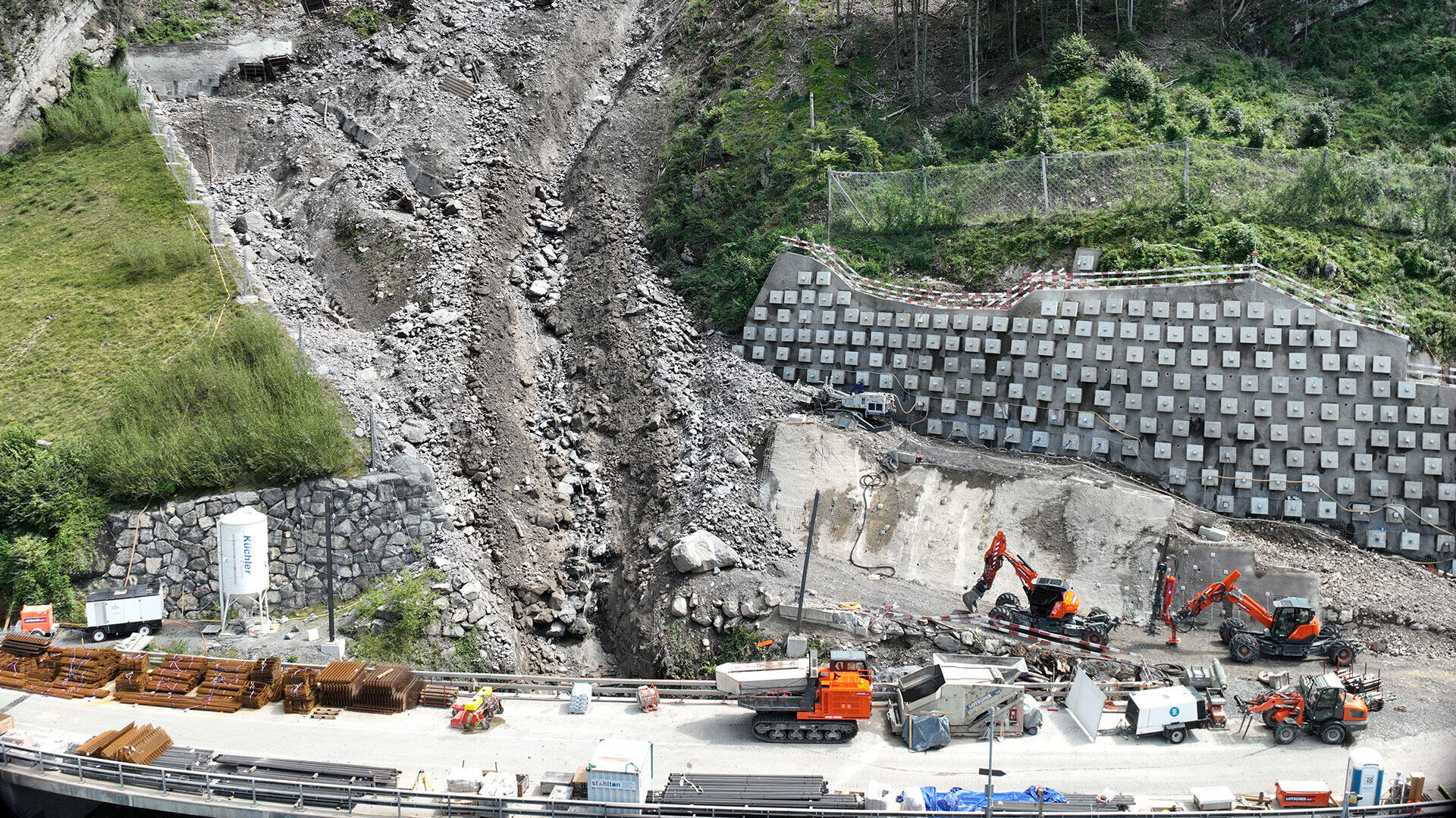 Ein Erdrutschgebiet mit Steinen und Schutt fließt einen Hang hinunter in Richtung einer Straße. Mehrere Baufahrzeuge und Arbeiter räumen und reparieren das Gelände in der Nähe einer Stützmauer und von Baumaterialien.