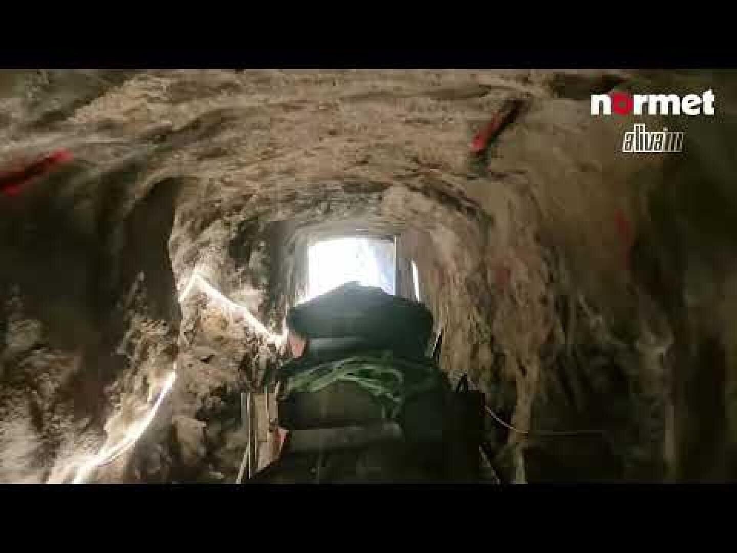 Blick aus dem Inneren eines felsigen Tunnels in Richtung des hellen Tunnelausgangs. Im Vordergrund sind Maschinen und Geräte zu sehen. Die Worte Normet und Aliva erscheinen in der oberen rechten Ecke.