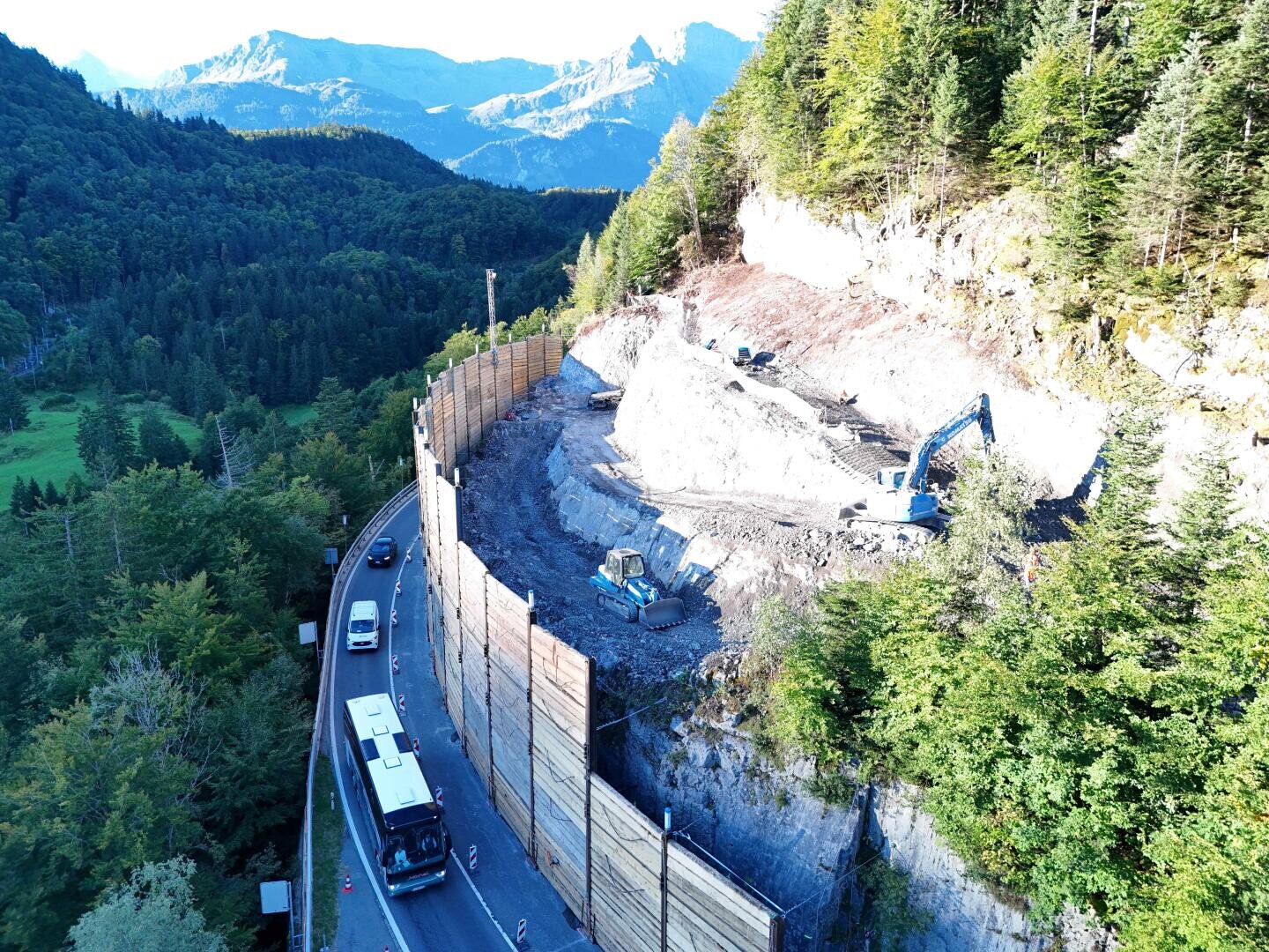 Eine Baustelle an einem steilen Hang neben einer kurvenreichen, von Fahrzeugen gesäumten Straße, mit einem blauen Bagger bei der Arbeit und hölzernen Stützmauern, die den Hang stützen, umgeben von dichtem Wald und fernen Bergen.