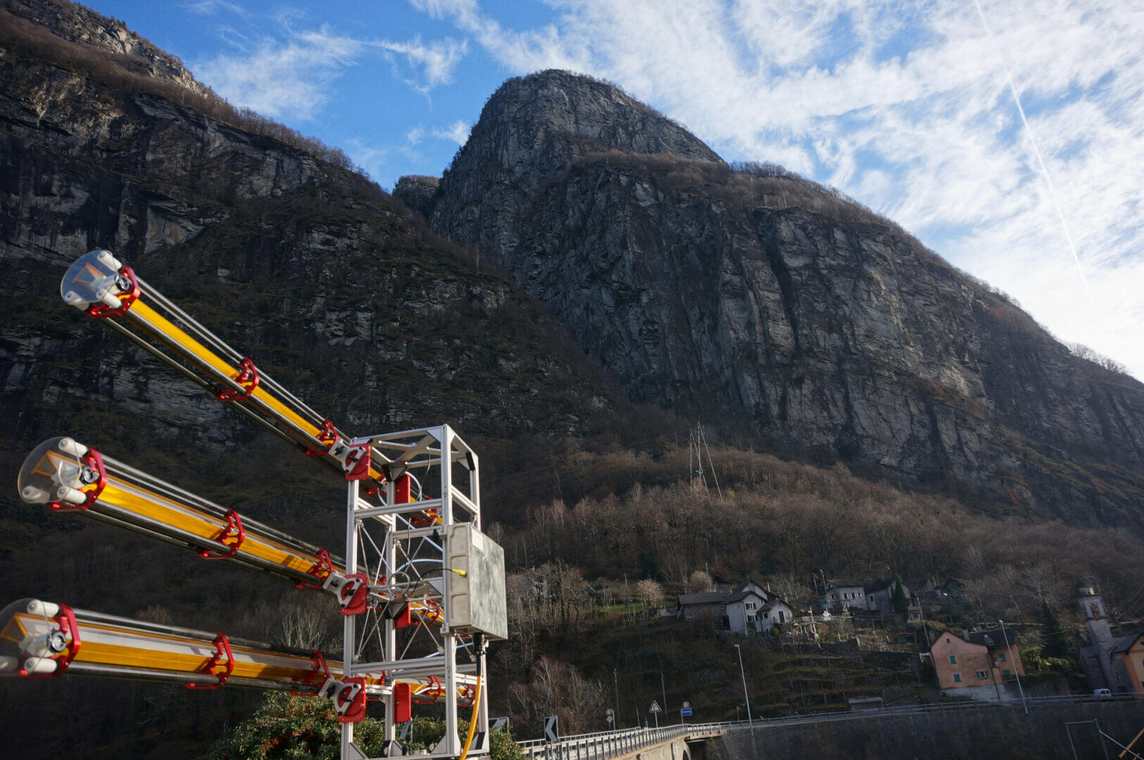 Ein großes Industriegebäude mit vier gelben und roten Röhren steht vor einem steilen, felsigen Berg mit vereinzelten Bäumen und ein paar kleinen Häusern an seinem Fuße, unter einem teilweise bewölkten blauen Himmel.