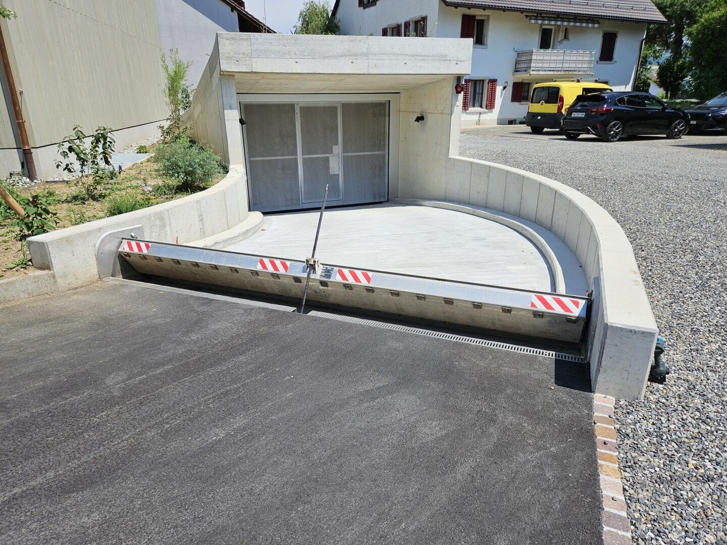 Eine moderne Garage mit einer geschwungenen Betoneinfahrt und einer versenkbaren Schranke an der Einfahrt; die Einfahrt ist glatt, und die Autos sind draußen geparkt, mit Häusern im Hintergrund.