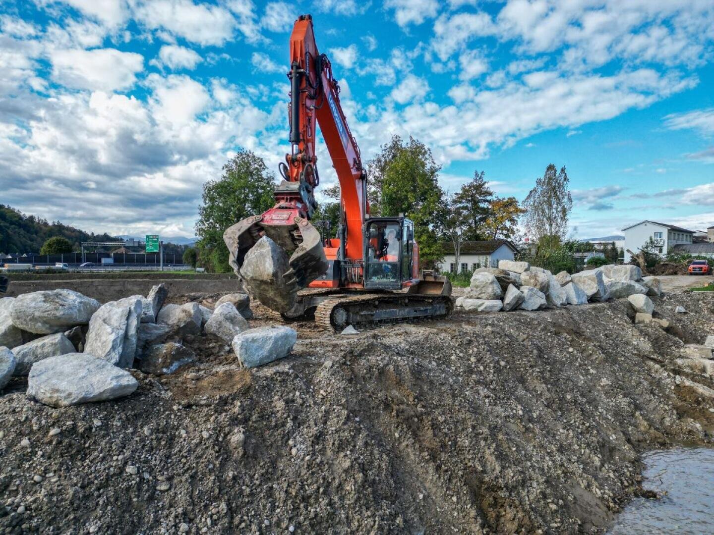 Ein orangefarbener Bagger hebt einen großen Felsen auf einer Baustelle an, die von Erde, Kies und anderen Felsen umgeben ist, mit Bäumen, Häusern und einem bewölkten Himmel im Hintergrund.