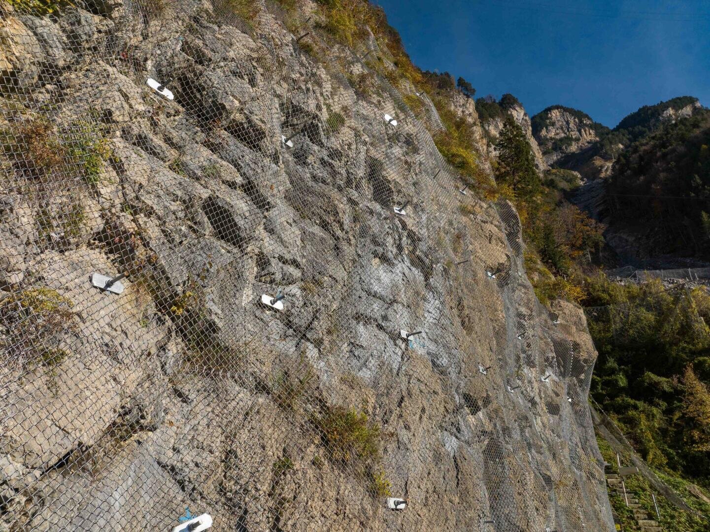 Eine steile Felswand, die mit Maschendraht und Metallankern zum Schutz vor Erdrutschen bedeckt ist, mit spärlicher Vegetation am Hang und Bergen im Hintergrund unter einem klaren blauen Himmel.