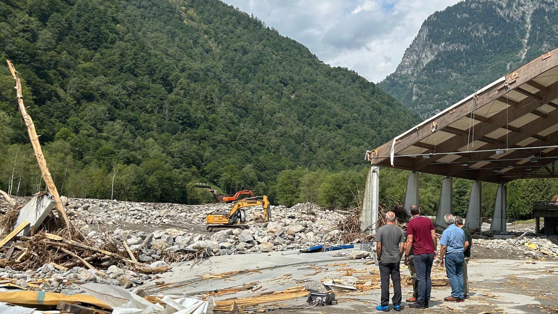 Vier Personen stehen in der Nähe eines teilweise zerstörten Gebäudes und beobachten zwei Bagger, die in einer bergigen, bewaldeten Gegend bei bewölktem Himmel zwischen Felsen und Trümmern arbeiten.