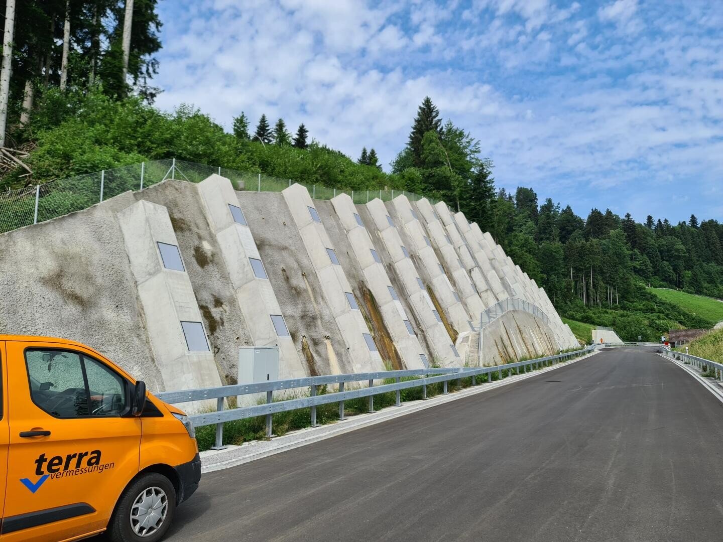 Ein leuchtend orangefarbener Lieferwagen mit Terravermessungen parkte neben einer gepflasterten Straße, neben einem steilen, mit großen, schrägen Betonplatten und kleinen Fenstern bewehrten Hang; grüne Bäume und Büsche im Hintergrund unter einem teilweise bewölkten Himmel.