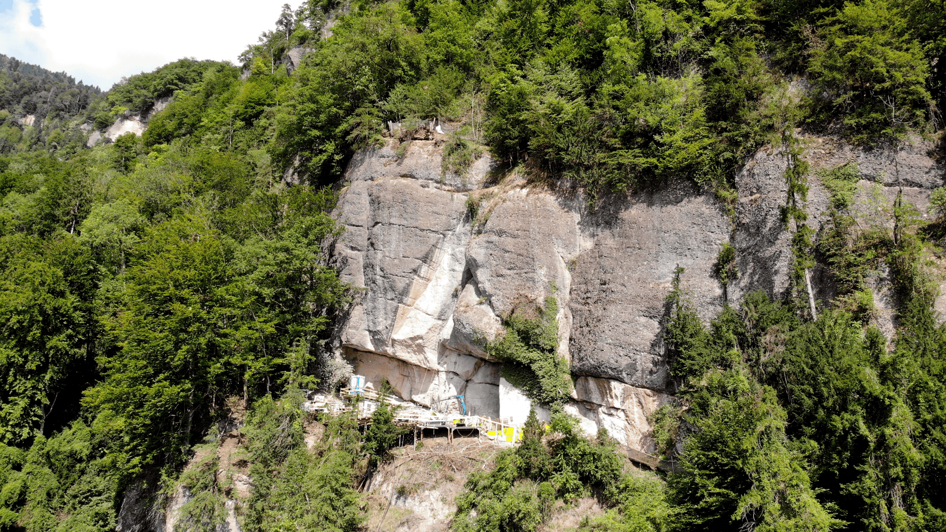 Eine kleine, farbenfrohe Struktur, die in einen felsigen Abhang gebaut wurde, umgeben von dichten grünen Bäumen und Vegetation, mit steil aufragenden Felswänden dahinter.