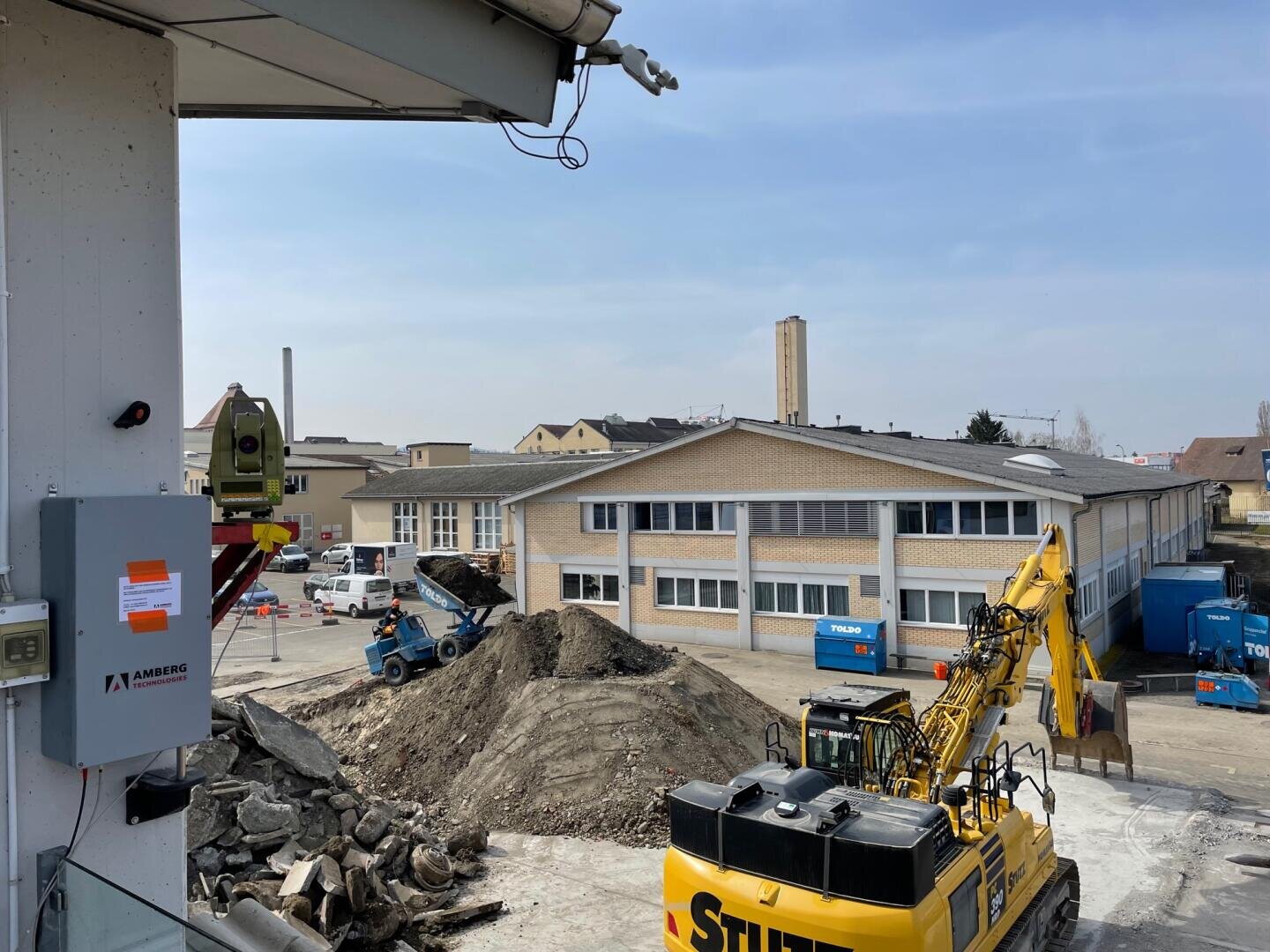 Eine Baustelle mit schweren Maschinen, darunter ein gelber Bagger und ein blauer Lader, die Erde und Schutt bewegen. Ein großer Erdhaufen liegt an einem klaren Tag vor einem beigen Industriegebäude.