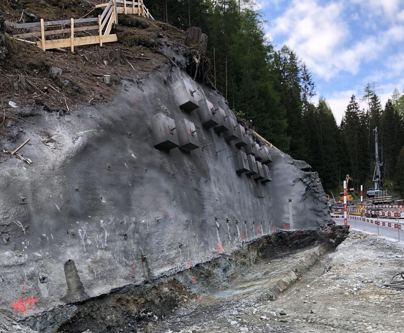Ein steiler, mit Beton und Metallstäben bewehrter Felshang steht neben einer im Bau befindlichen Straße, die mit roter Farbe markiert ist. Oben befinden sich Holzzäune und Bäume, und auf der rechten Seite säumen Sicherheitsbarrieren die Straße.