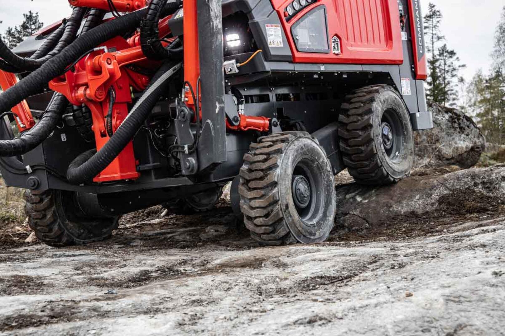 Nahaufnahme eines roten Geländewagens mit großen, robusten Reifen, der über unebenes, felsiges Gelände in einem bewaldeten Außenbereich fährt.