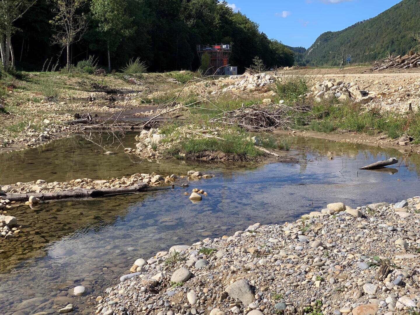 Ein flacher, felsiger Bach fließt durch ein trockenes, steiniges Flussbett, umgeben von grünen Bäumen und Hügeln unter einem blauen Himmel. An den Ufern liegen Treibholz und Äste verstreut.