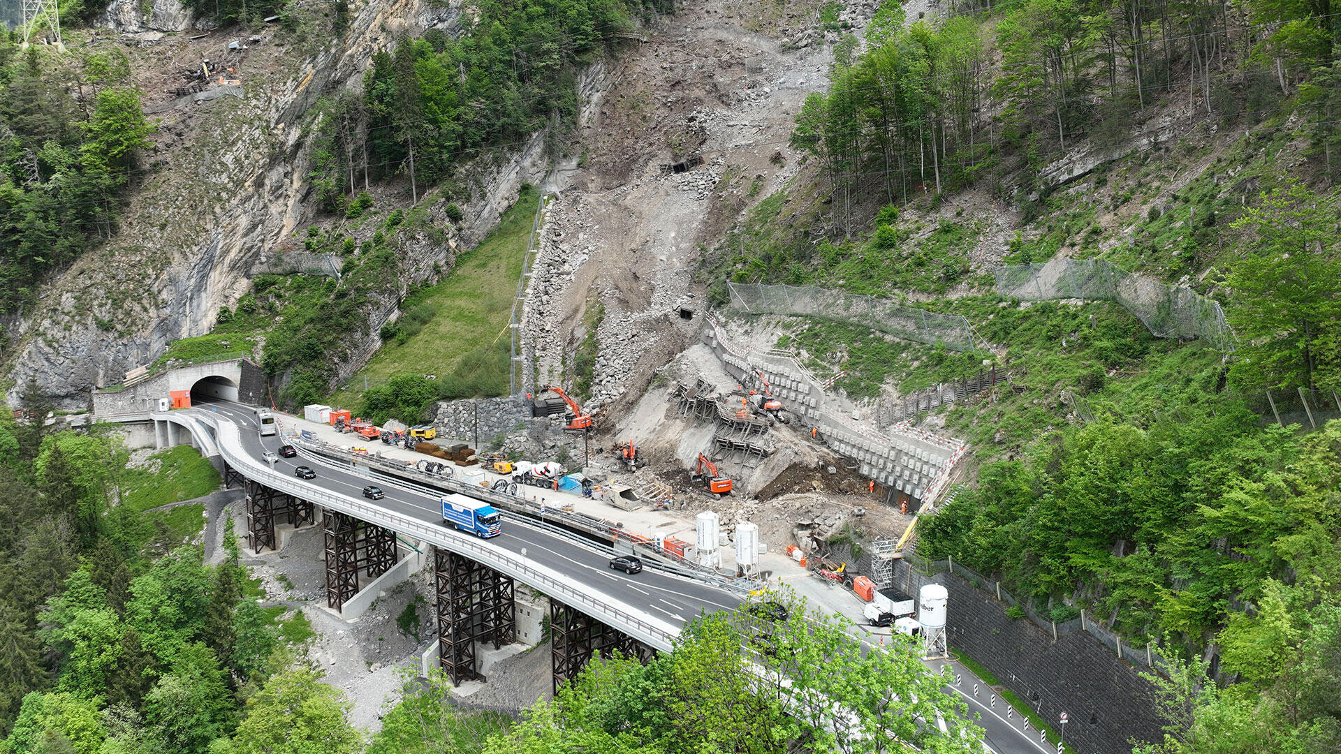 Eine Bergstraße und eine Brücke werden derzeit repariert. In der Nähe eines Tunneleingangs sind Baufahrzeuge und Arbeiter zu sehen. Das Gebiet ist von Bäumen und felsigen Hängen umgeben. Auf einem Teil der Straße herrscht weiterhin Verkehr.