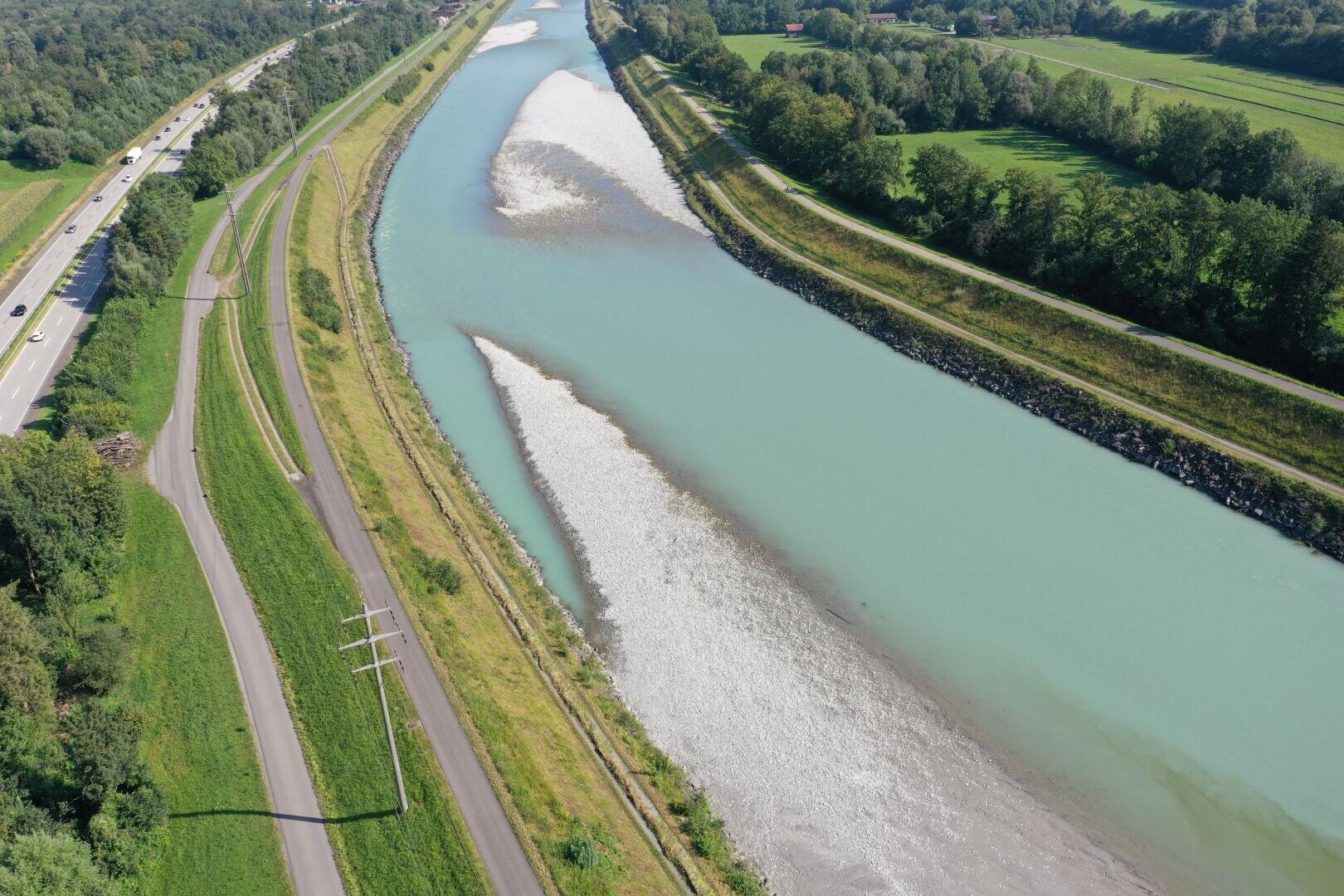 Luftaufnahme eines gewundenen Flusses mit hellblauem Wasser, getrennt durch eine Kiesbank. Der Fluss wird von grünen Feldern, Bäumen und parallelen Straßen auf beiden Seiten flankiert. Auf einer Straße auf der linken Seite sind Autos zu sehen.