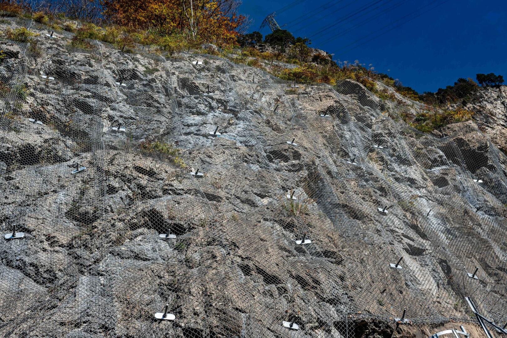 Ein steiler Felshang ist mit Maschendraht bedeckt und mit Metallankern gesichert, um Steinschlag zu verhindern. Auf dem Gipfel und am Hang wächst etwas Vegetation. Der Himmel darüber ist klar und blau.