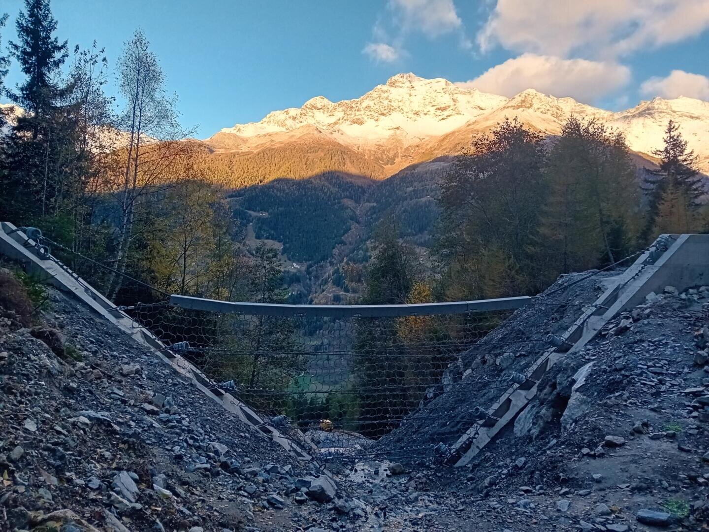 Eine Absperrung aus Draht und Beton erstreckt sich über eine felsige Lücke in einem Waldgebiet. Im Hintergrund sind schneebedeckte Berge und Bäume zu sehen, die bei teilweise bewölktem Himmel vom Sonnenlicht angestrahlt werden.