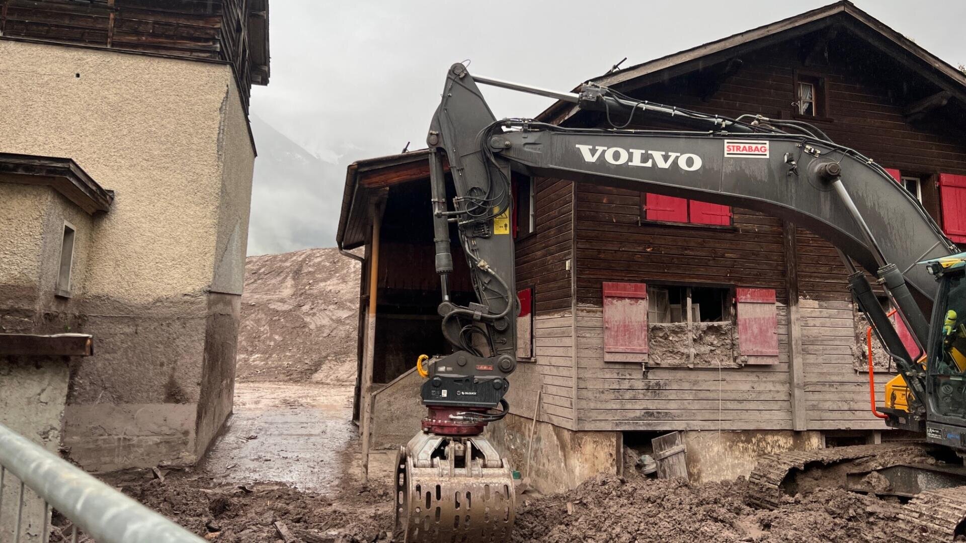 Ein Volvo-Bagger räumt nach einem Erdrutsch Schlamm vor Holz- und Betonhäusern weg, wobei der Schlamm den Boden und die Hauswände bedeckt. Der Himmel ist bedeckt, und das Gebiet scheint stark beschädigt zu sein.