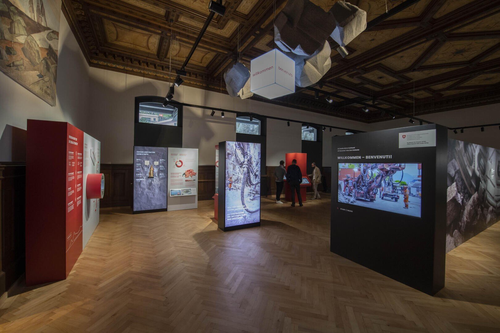 Ausstellungshalle mit Holzdecke, interaktiven Anzeigetafeln, Wandbildschirmen und Besuchern, die Exponate über Katastrophenschutz und Notfalldienste betrachten. Warme Beleuchtung unterstreicht die informativen Displays.