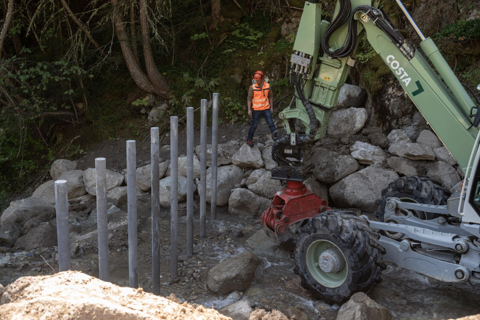 Ein Bauarbeiter in orangefarbener Weste und mit Helm steht in der Nähe eines felsigen Baches, während ein grüner Bagger Metallpfosten entlang des Gewässerrandes aufstellt. Die Baustelle ist von Bäumen und großen Felsen umgeben.
