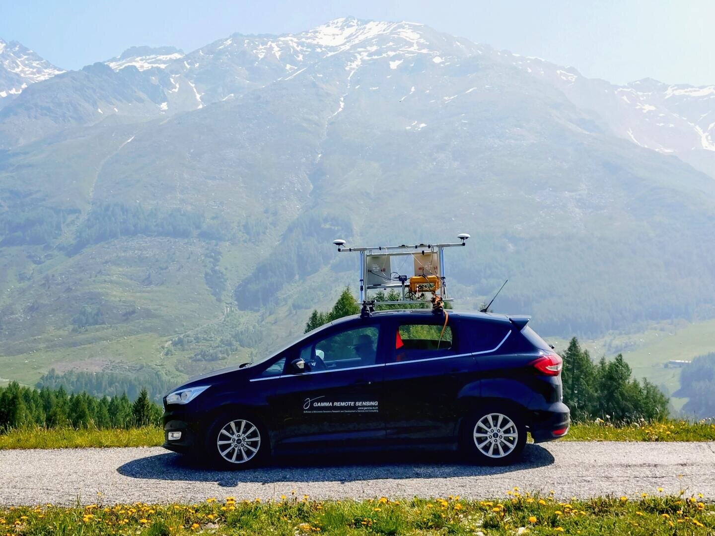 Ein schwarzes Auto mit wissenschaftlichen Instrumenten auf dem Dach ist auf einer Straße geparkt, mit grünen Feldern und hohen Bergen im Hintergrund unter einem klaren Himmel.