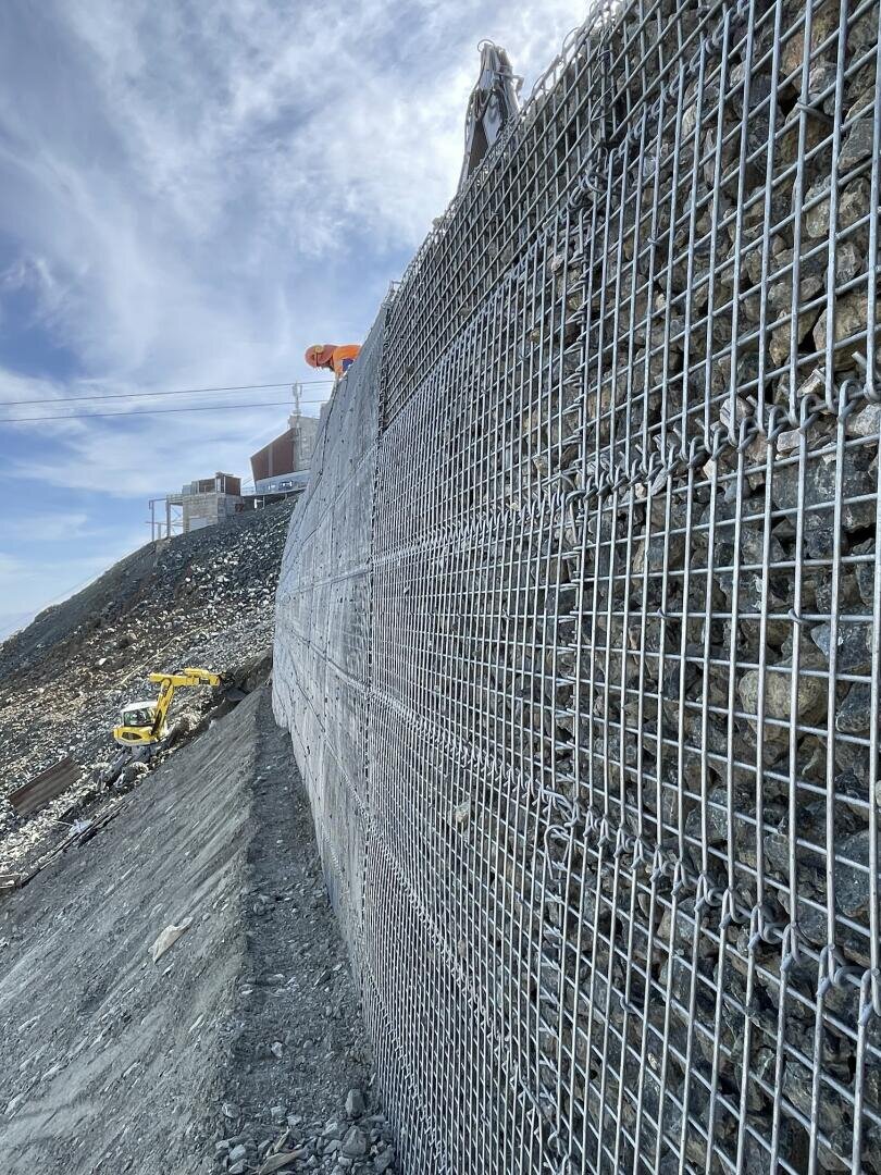 Nahaufnahme einer mit Steinen gefüllten Stützmauer aus Drahtgeflecht auf einer abschüssigen Baustelle. Im Hintergrund sind Baumaschinen und Gebäude bei teilweise bewölktem Himmel zu sehen.
