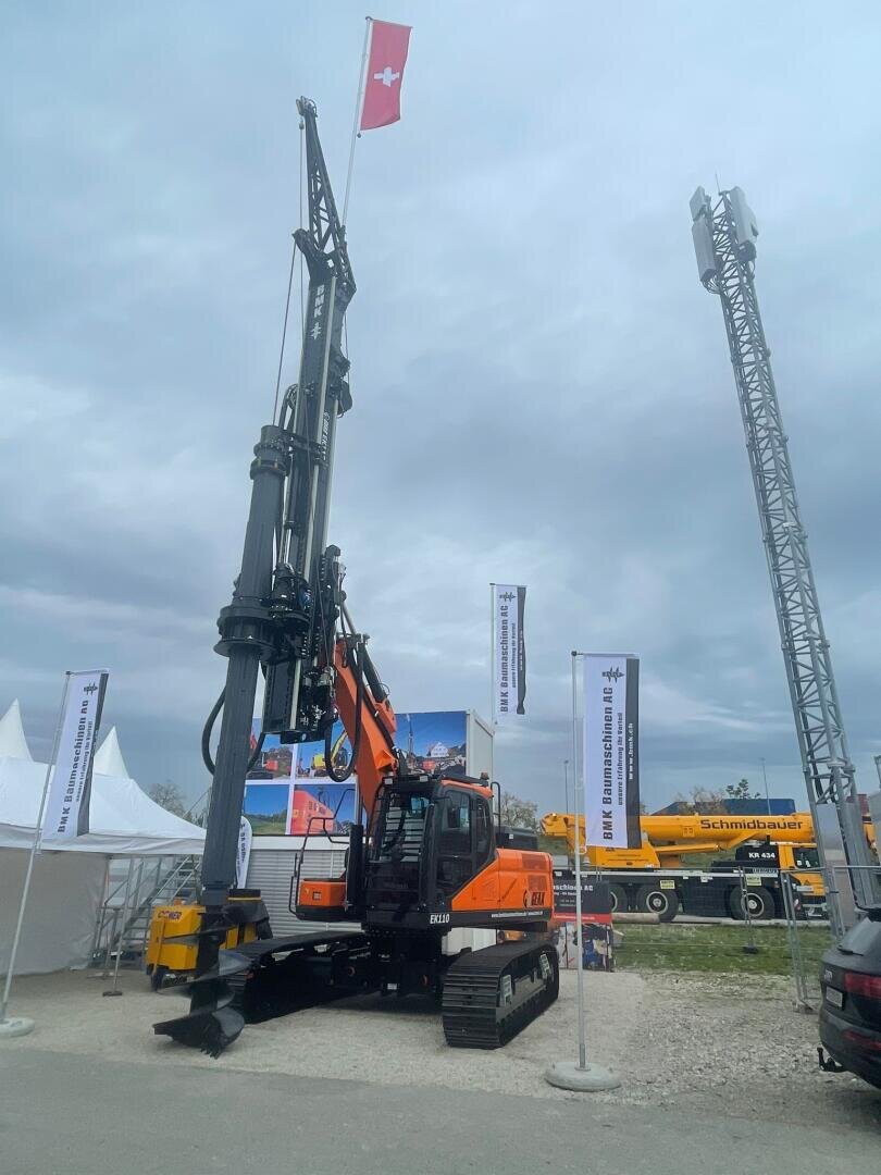 Eine große orangefarbene Bohrmaschine steht im Freien unter einem wolkenverhangenen Himmel, über der eine Schweizer Flagge an einem hohen Mast gehisst ist. In der Nähe stehen Zelte, Transparente und ein weiteres gelbes Baufahrzeug.