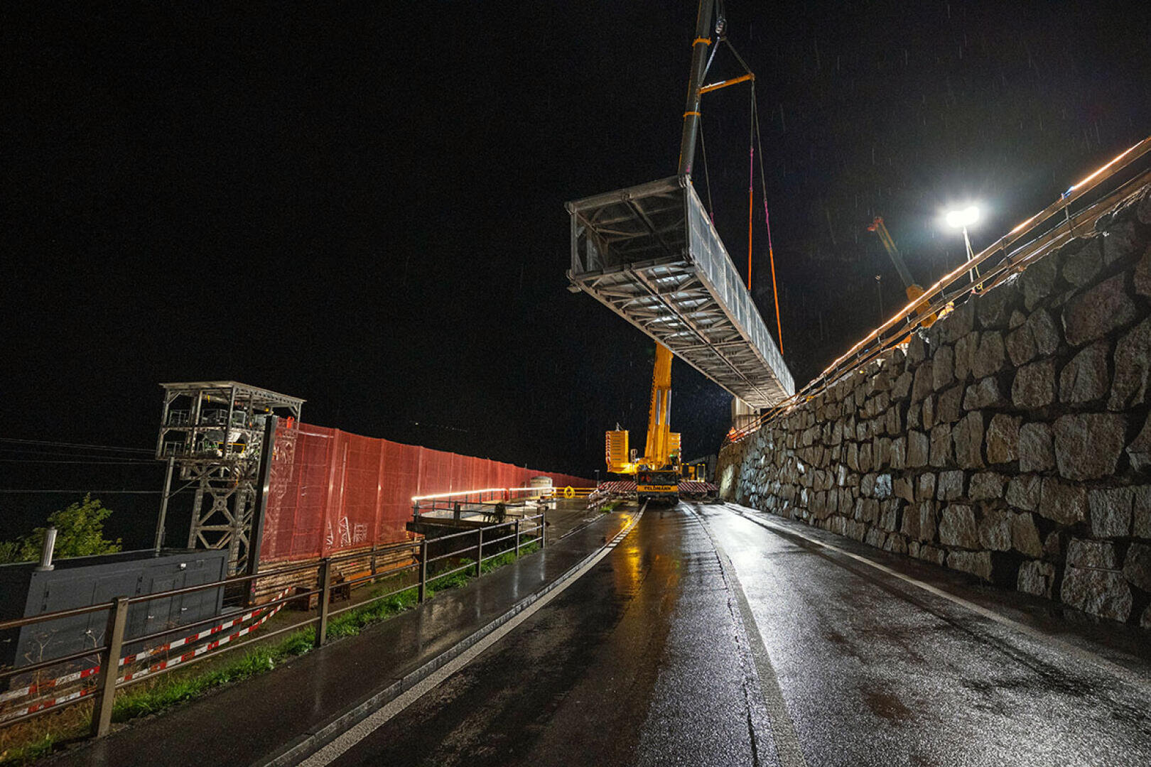 Ein großes Brückenteil aus Metall wird nachts von einem Kran angehoben. Die nasse, spiegelnde Straße, die Steinmauer und die Bauabsperrungen sind hell erleuchtet.