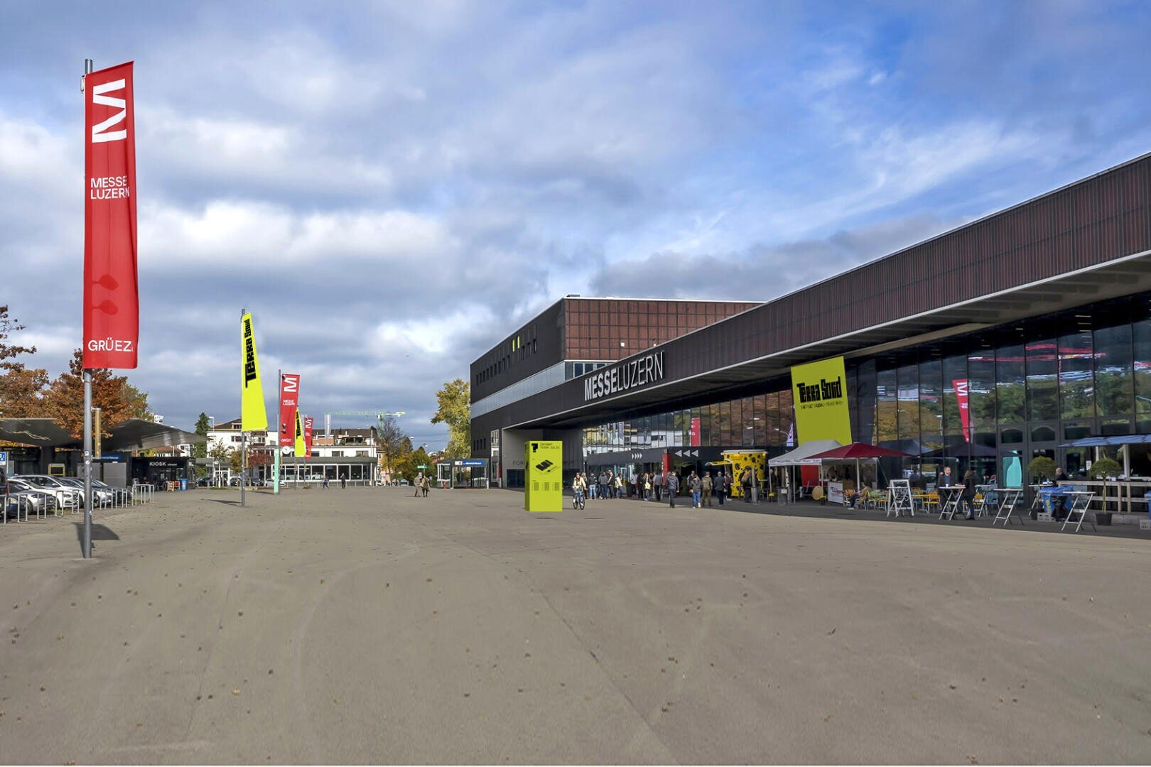 Weite Sicht auf das Messe Luzern Kongresszentrum mit roten Fahnen, Sitzgelegenheiten im Freien und Menschen in der Nähe des Eingangs unter einem teilweise bewölkten Himmel. Der Platz ist weitläufig, mit heller Beschilderung und Bäumen auf der linken Seite.