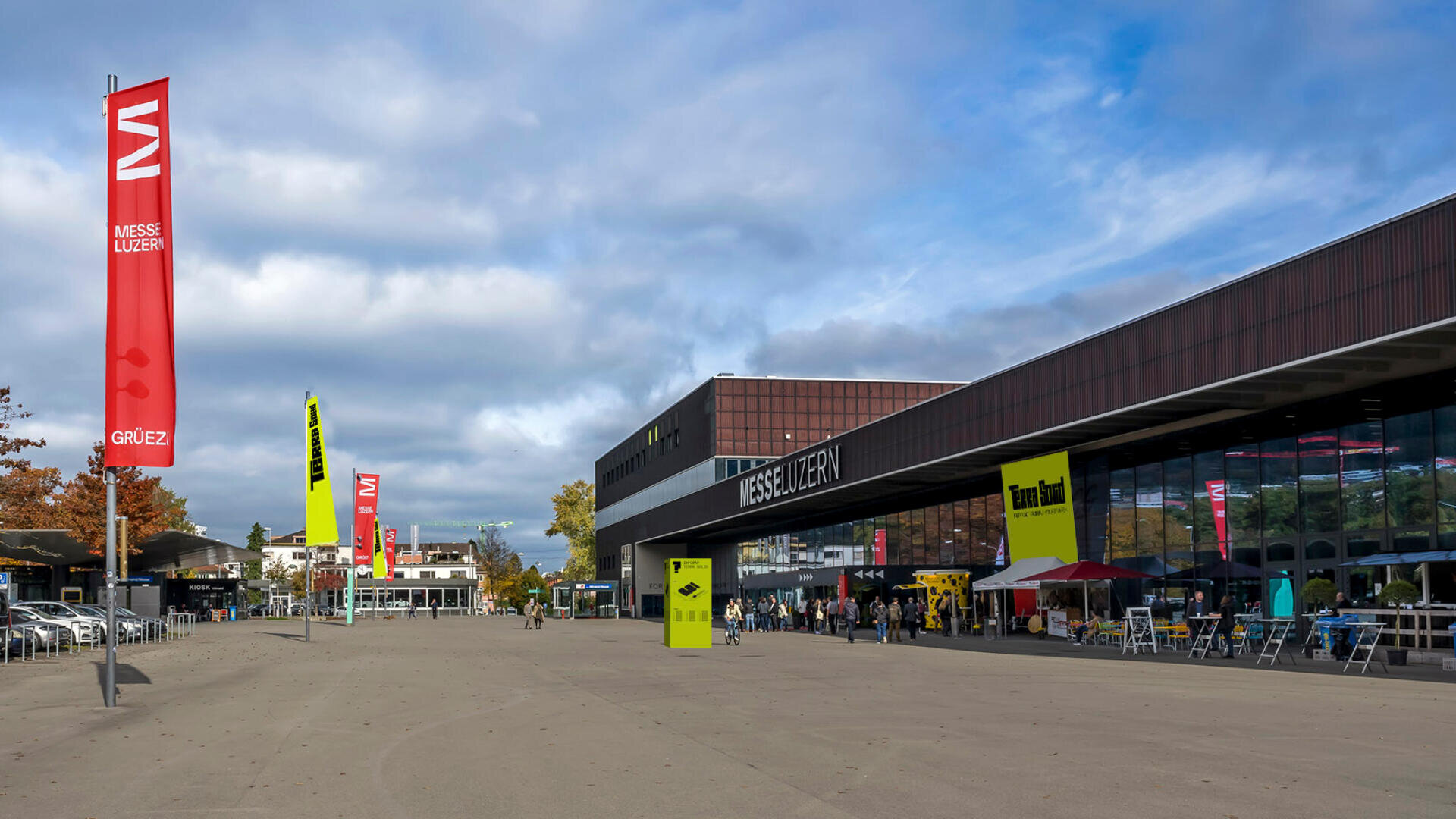 Breiter Platz vor dem Kongresszentrum Messe Luzern mit roten und gelben Bannern, Menschen, die spazieren gehen oder an Tischen sitzen, und darüber ein teilweise bewölkter Himmel.