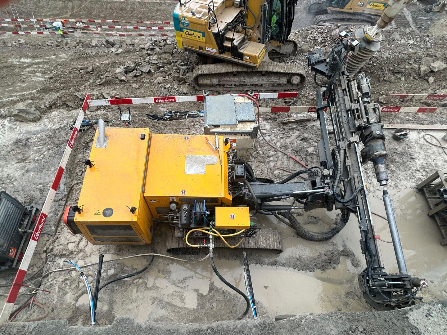 Eine gelbe Bohrmaschine mit Raupen arbeitet auf einer schlammigen Baustelle, umgeben von roten und weißen Sicherheitsbarrieren. Ein weiterer Bagger ist in der Nähe zu sehen, zusammen mit verstreutem Baumaterial.