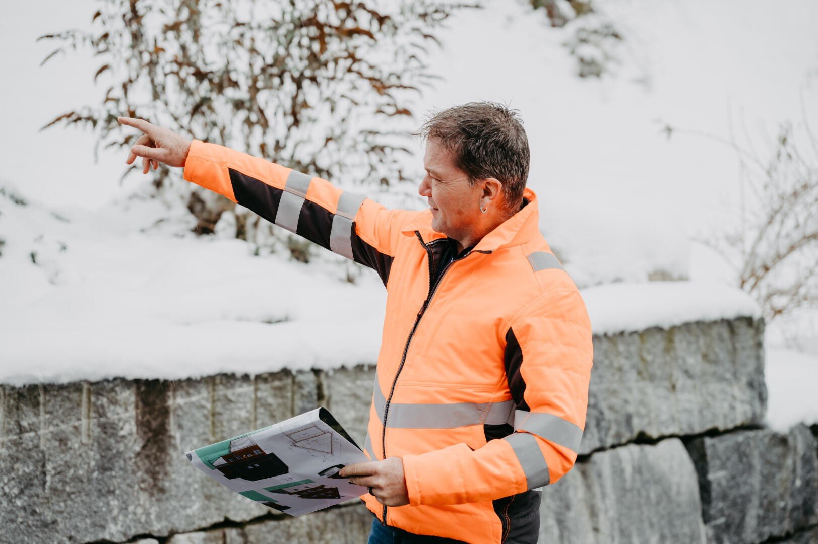 Ein Mann in einer leuchtend orangefarbenen, reflektierenden Jacke steht draußen in einer verschneiten Umgebung, hält eine Karte oder ein Dokument in der Hand und zeigt nach links, um möglicherweise Anweisungen zu geben oder auf etwas in der Ferne hinzuweisen.