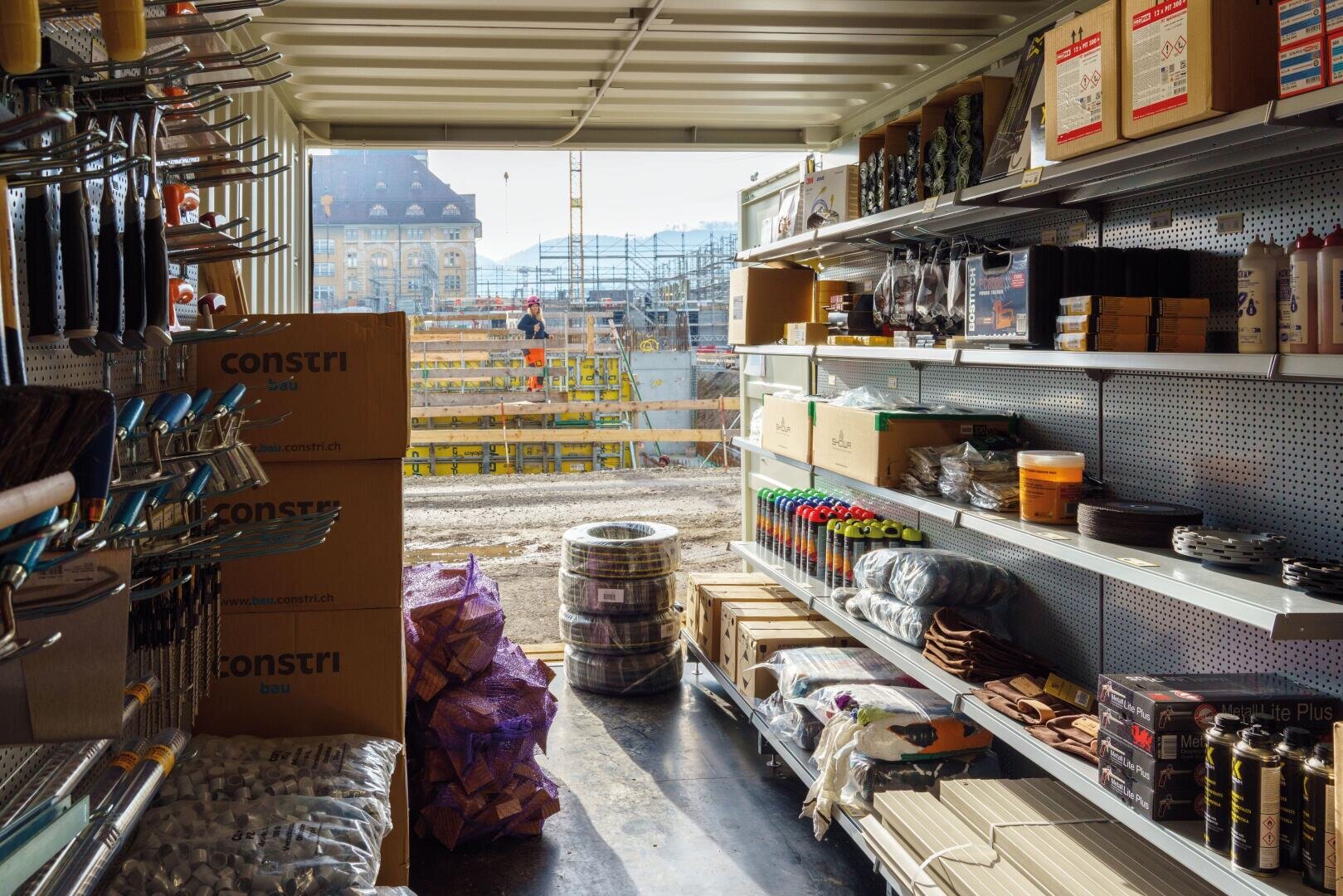 Ein Lagercontainer, gefüllt mit Bauwerkzeugen und Baumaterialien auf Regalen, mit Lichteinfall durch eine offene Tür mit Blick auf eine aktive Baustelle und Arbeiter im Hintergrund.