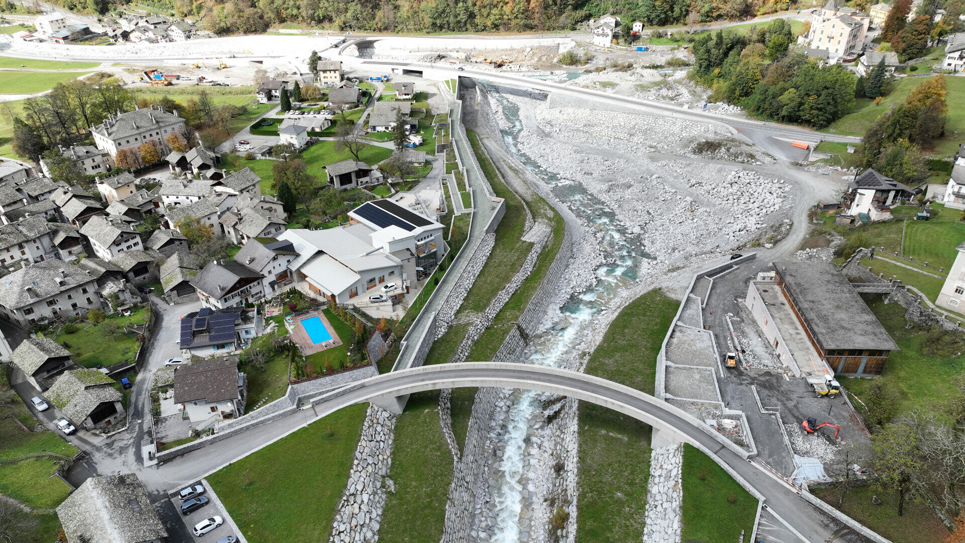 Luftaufnahme eines kleinen Dorfes mit Steinhäusern, einem modernen Haus mit Pool, einer geschwungenen Fußgängerbrücke über ein felsiges Flussbett und grünen Hügeln im Hintergrund. Eine Straße überquert eine weitere Brücke im oberen Bildbereich.