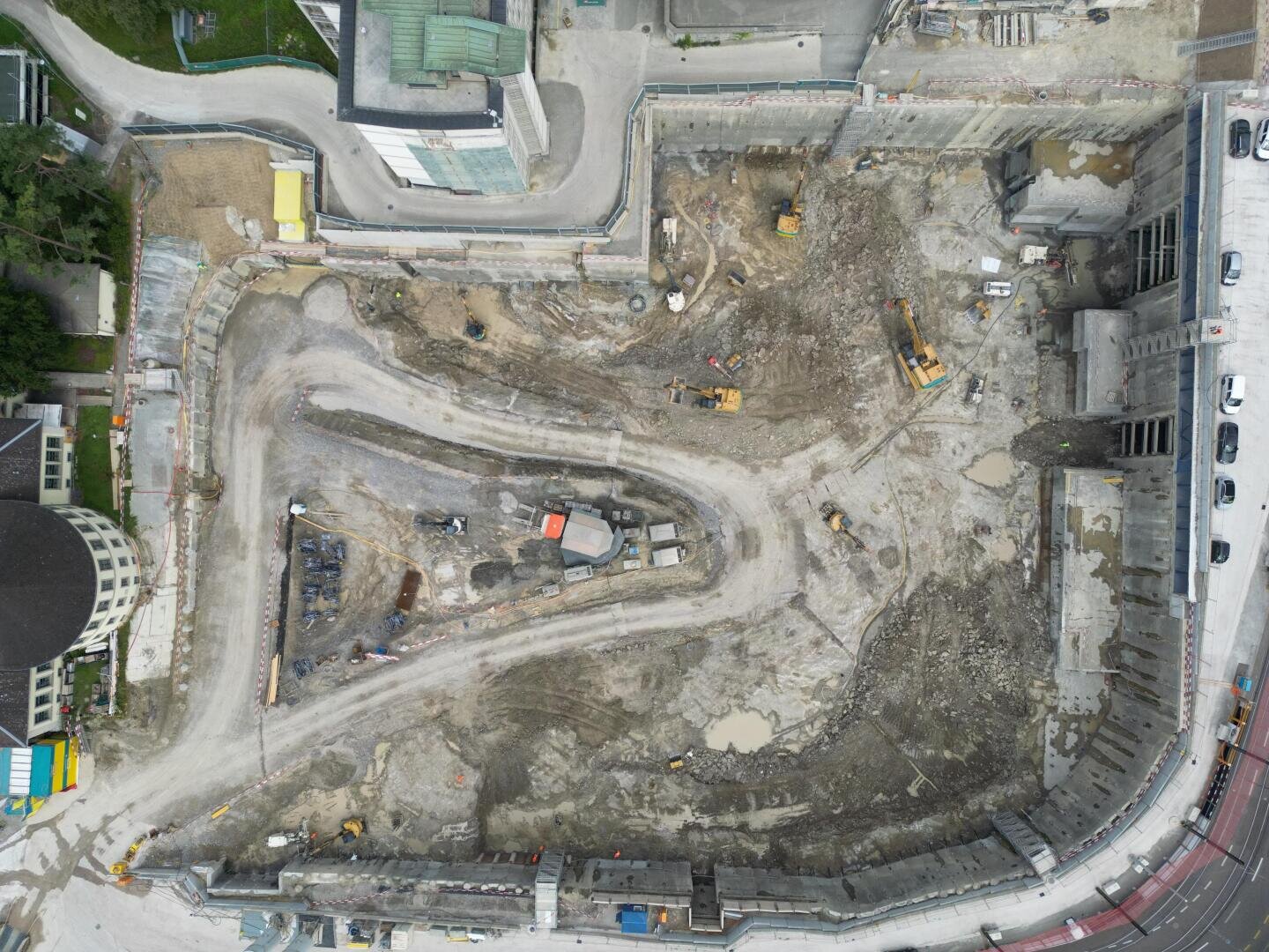 Luftaufnahme einer Großbaustelle mit Baggern, unbefestigten Wegen und Gebäudefundamenten. Umgeben von Straßen, geparkten Autos und teilweise fertiggestellten Gebäuden. Die Baustelle wirkt geschäftig und organisiert.