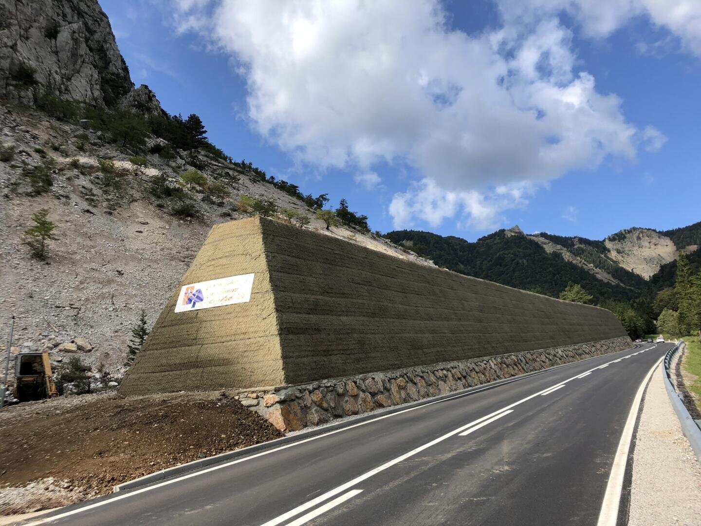 Eine hohe, schräge Stützmauer aus bewehrter Erde verläuft entlang einer asphaltierten Bergstraße, mit einem Bauschild an der Mauer und felsigen Hügeln im Hintergrund unter einem teilweise bewölkten Himmel.