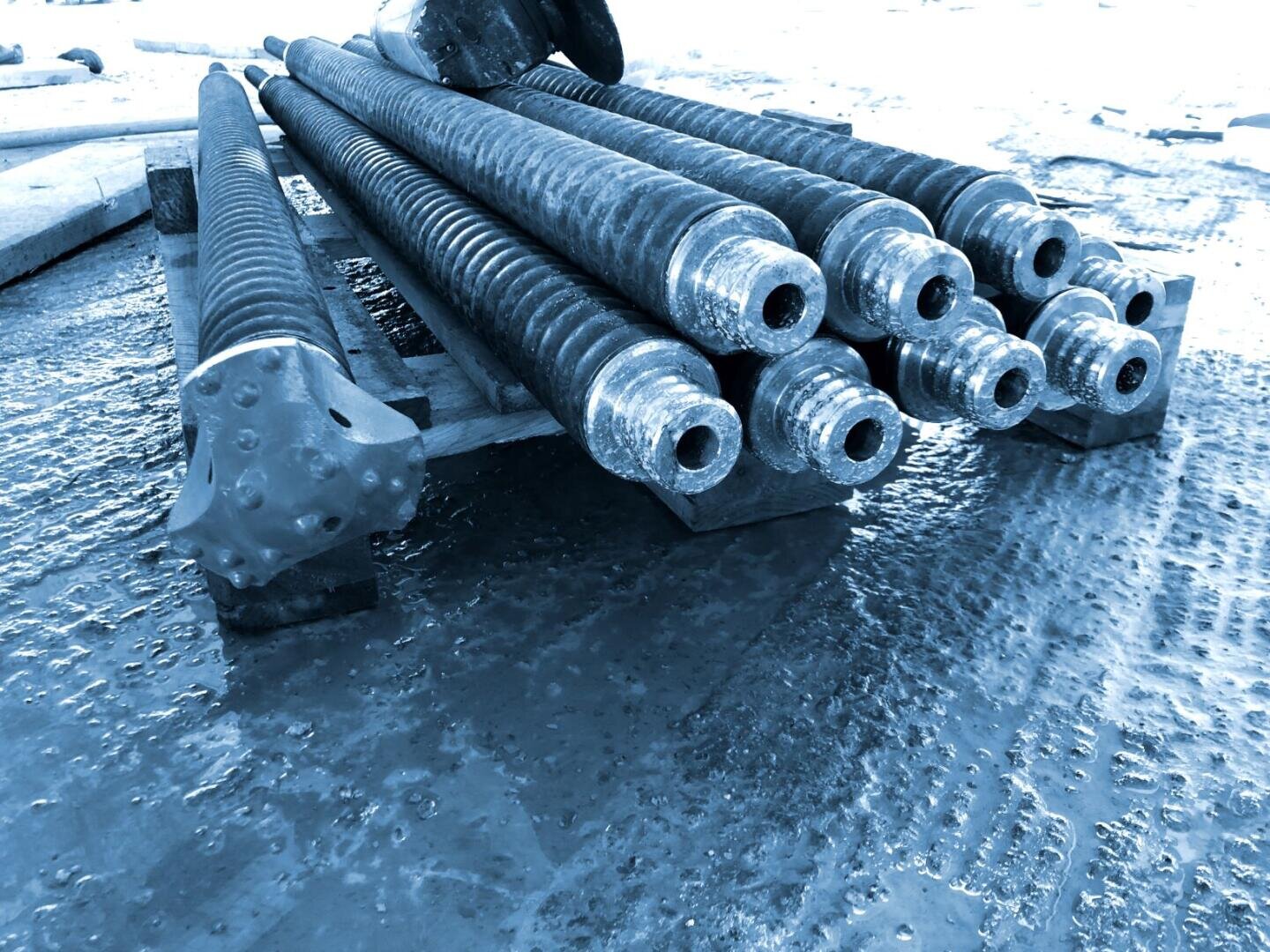 Mehrere große, schwere Metallbohrgestänge und ein Bohrer sind auf einer nassen, strukturierten Bodenoberfläche gestapelt, möglicherweise auf einer Industrie- oder Baustelle. Das Bild ist blau eingefärbt.