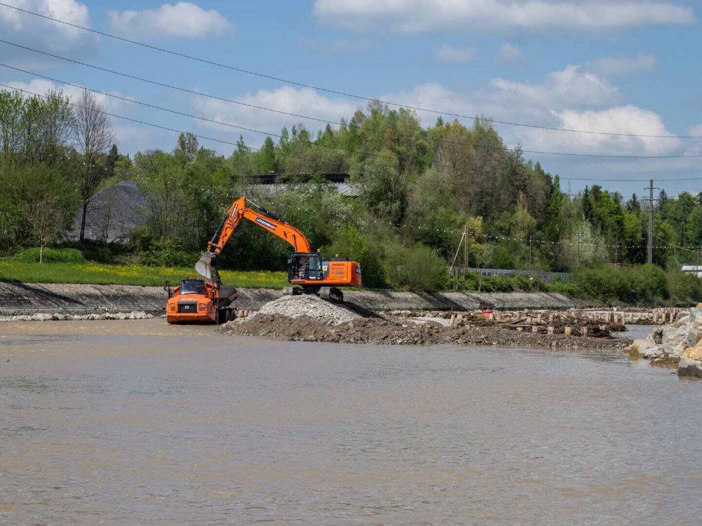 Ein orangefarbener Bagger lädt Erde auf einen Kipplaster neben einem Fluss, mit Bäumen und Grünpflanzen im Hintergrund unter einem teilweise bewölkten Himmel.