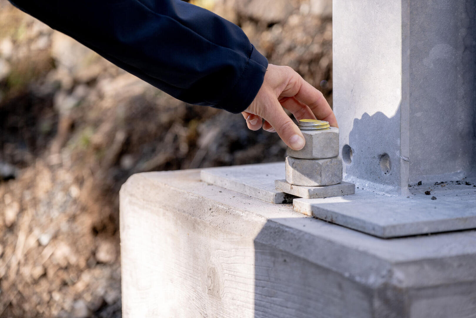 Die Hand einer Person verstellt große Metallmuttern an einem Bolzen am Fuß einer Metallkonstruktion, die im Freien auf einem Betonfundament verankert ist.