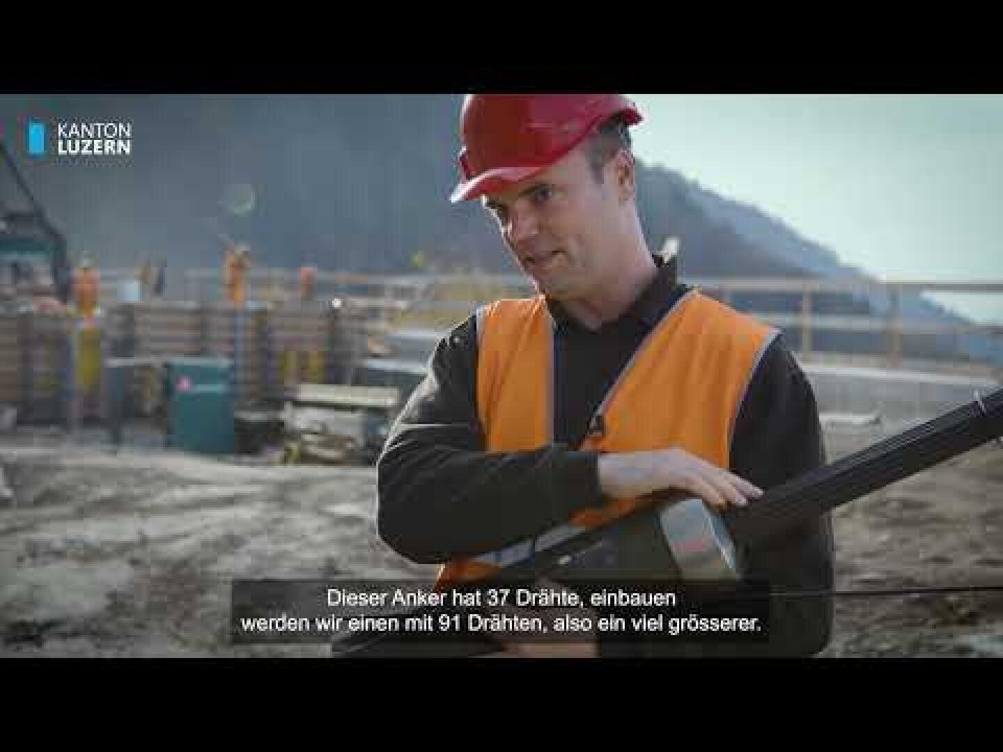 Ein Bauarbeiter in orangefarbener Weste und rotem Helm hält ein großes Werkzeug auf einer Baustelle. Im Hintergrund sind Berge zu sehen. Auf dem Bildschirm erscheinen deutsche Untertitel und ein "Kanton Luzern"-Logo.