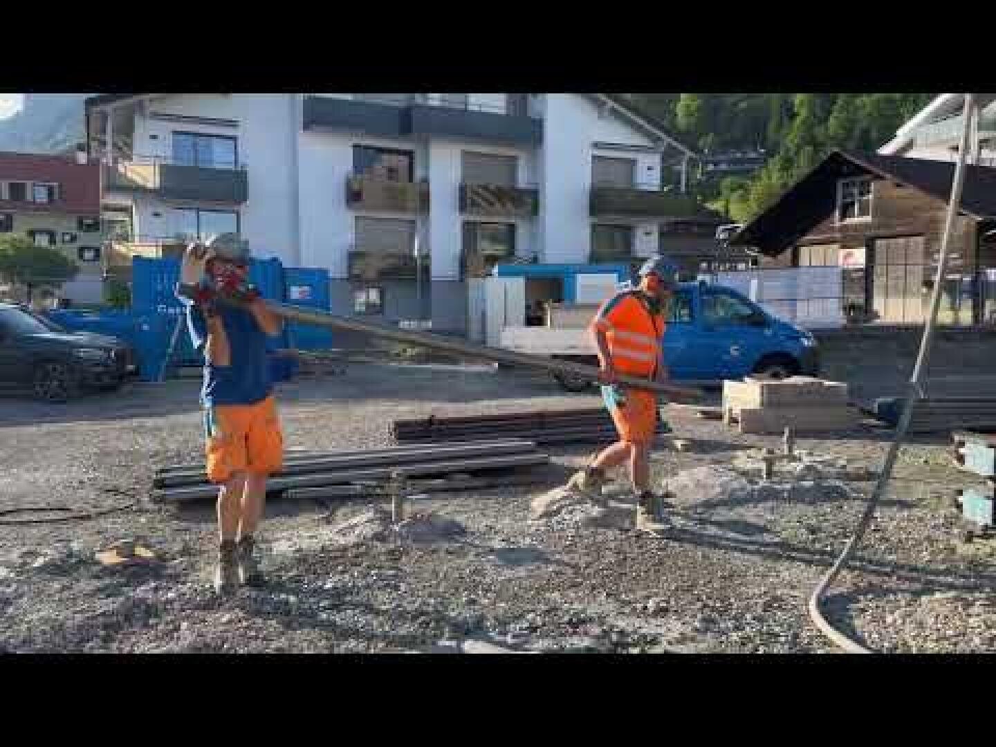 Zwei Bauarbeiter in Schutzkleidung tragen einen langen Metallträger auf einer Baustelle. Im Hintergrund sind Wohngebäude, Materialstapel und ein blauer Lieferwagen zu sehen.