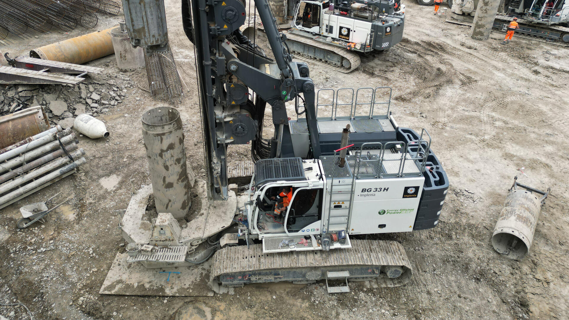 Eine große Bohrmaschine arbeitet auf einer Baustelle, umgeben von Betonrohren, Schutt und Arbeitern in orangefarbener Schutzkleidung. Eine weitere schwere Maschine und Baumaterialien sind im Hintergrund zu sehen.