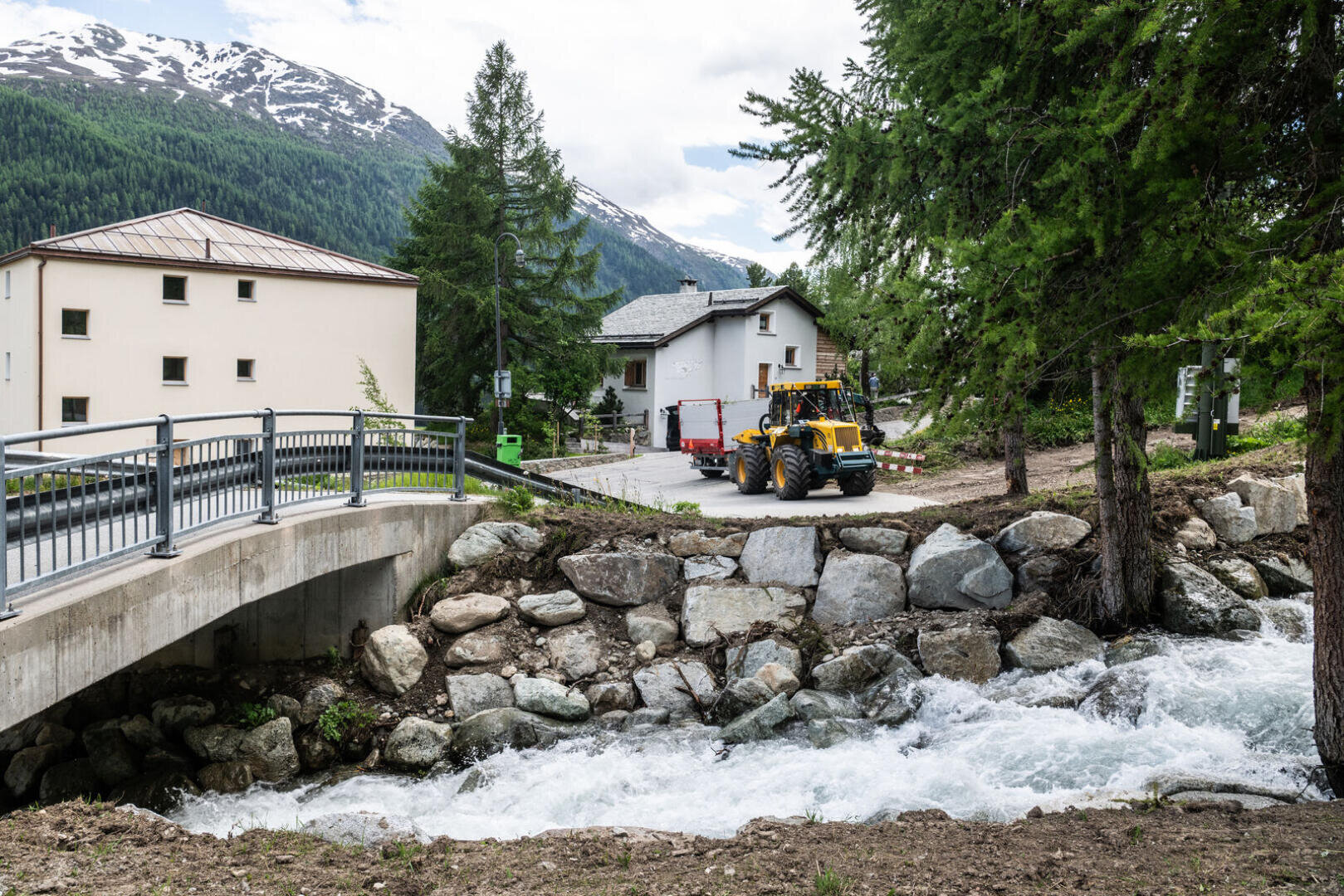 Ein kleiner Fluss fließt unter einer Betonbrücke über steinerne Dämme, ein gelber Traktor und ein roter Anhänger sind in der Nähe von weißen Häusern und Kiefern geparkt, vor dem Hintergrund bewaldeter Berge und eines bewölkten Himmels.