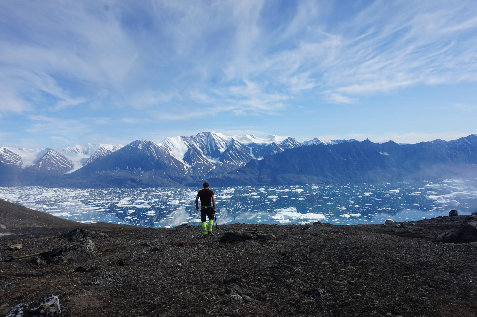 Eine Person steht auf felsigem Grund und blickt auf einen weiten, eisigen Fjord voller schwimmender Eisberge, mit schneebedeckten Bergen und einem blauen, wolkenverhangenen Himmel im Hintergrund.