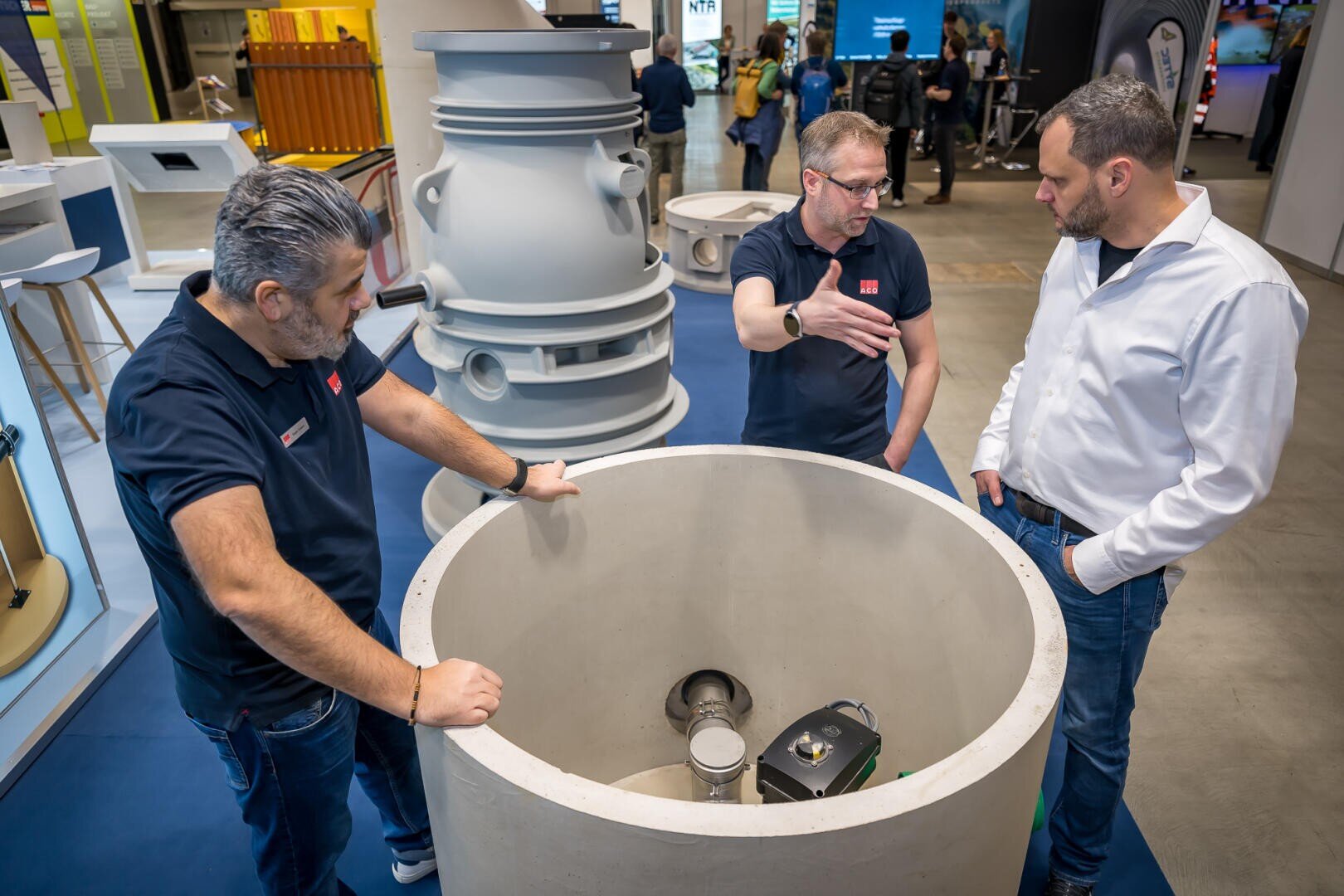 Drei Männer stehen auf einer Ausstellung um einen großen Industriezylinder aus Beton herum und diskutieren über dessen Eigenschaften. Zwei tragen marineblaue Hemden, einer ein weißes Hemd. Im Hintergrund sind Industrieanlagen und Displays zu sehen.