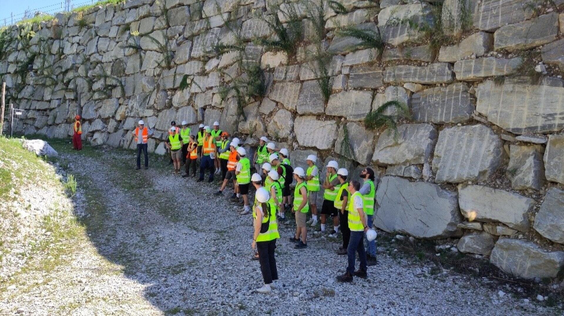 Eine Gruppe von Personen, die Sicherheitswesten und Schutzhelme tragen, steht im Freien neben einer großen Steinmauer und hört einer Person in roter Kleidung zu, möglicherweise während einer Baustelleninspektion oder -besichtigung.