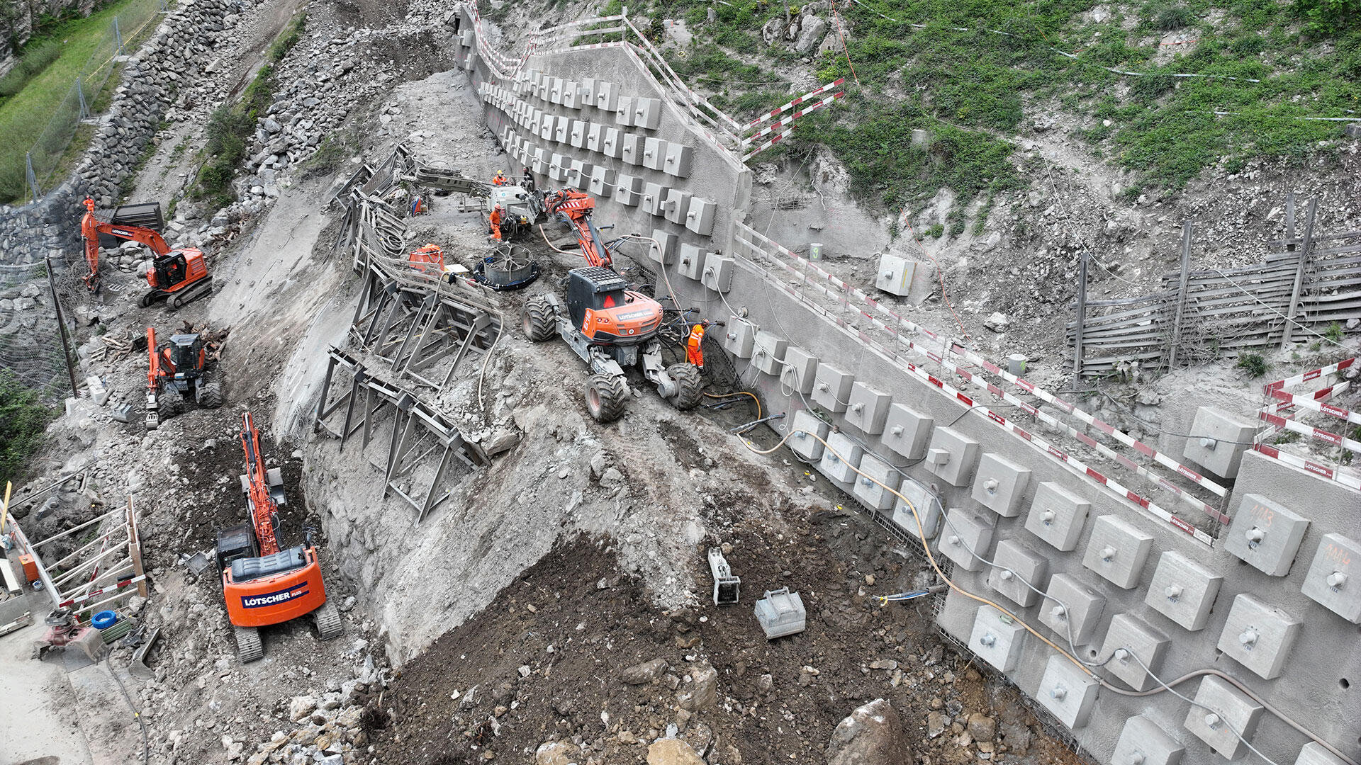 Eine Baustelle am Hang mit mehreren orangefarbenen Baggern und Arbeitern, die einen steilen Hang mit Betonkonstruktionen verstärken. Das Gelände ist von Erde, Steinen und provisorischen Zäunen umgeben.