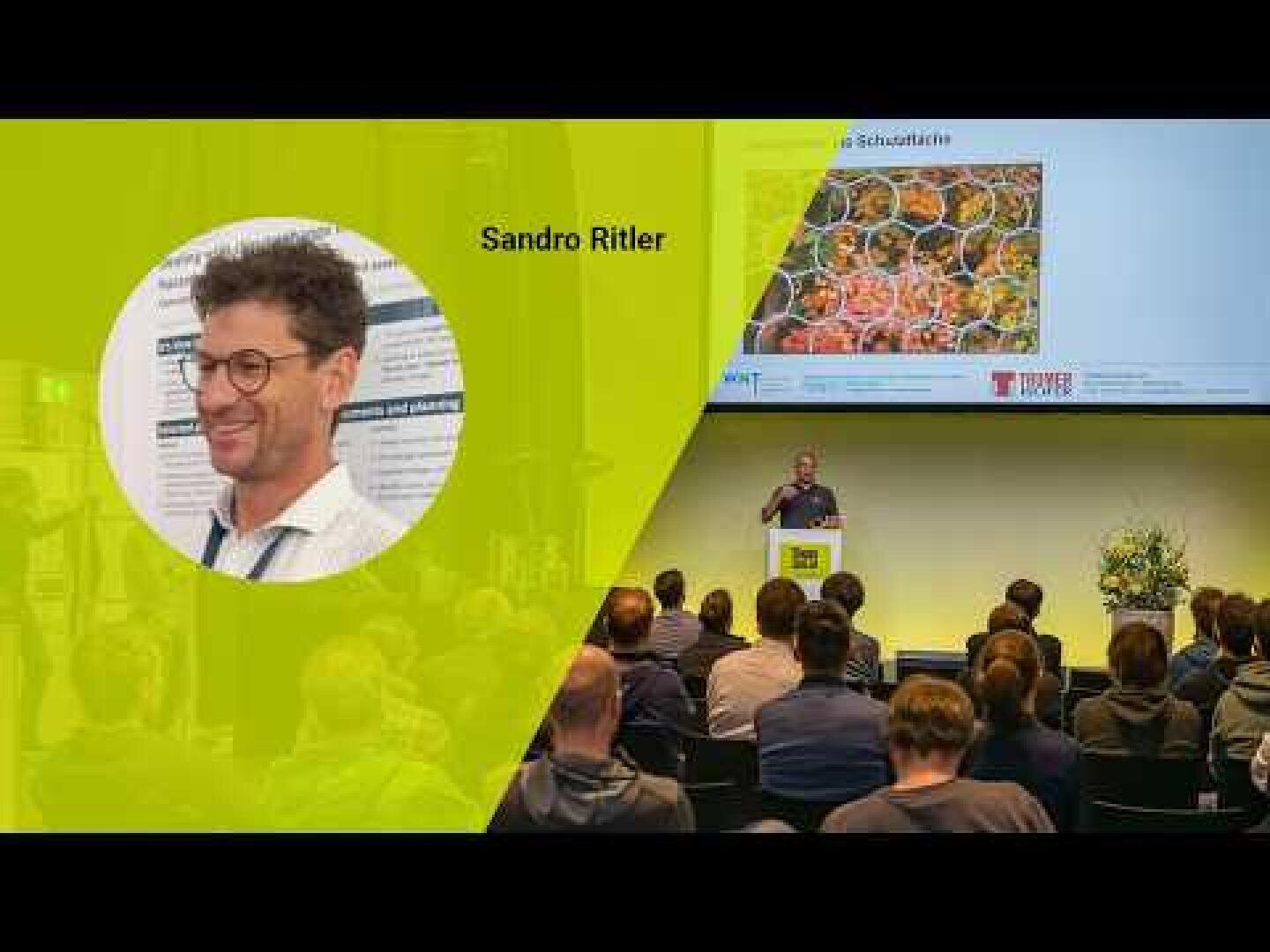 In einem Hörsaal findet eine Präsentation statt. Auf einem Dia ist das Foto eines Mannes mit der Aufschrift Sandro Ritler auf gelbem Hintergrund zu sehen. Das Publikum hört zu, während ein Redner vor einem projizierten Bild spricht.