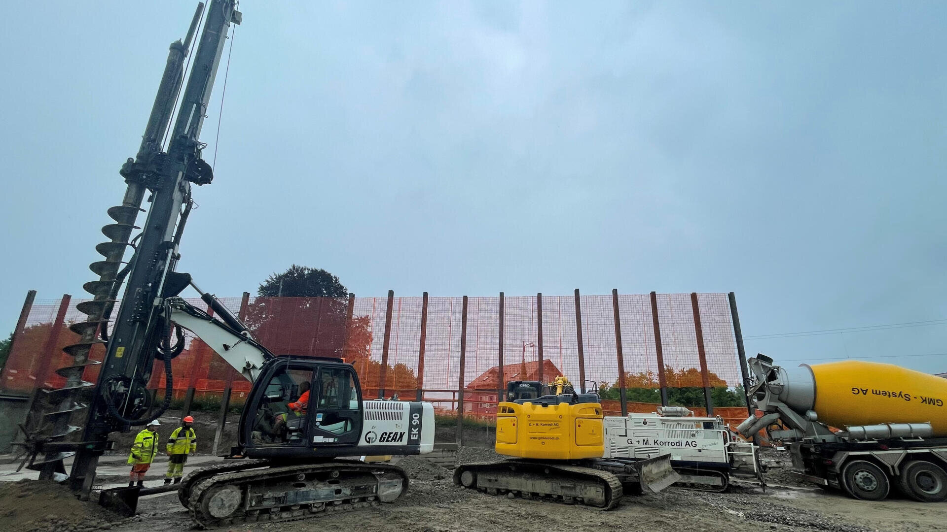 Baustelle mit großen Bohr- und Baggermaschinen, Arbeitern in Schutzausrüstung und einem gelben Zementmischer-LKW. Im Hintergrund sind bei bewölktem Himmel eine rote Gitterabsperrung und Gebäude zu sehen.