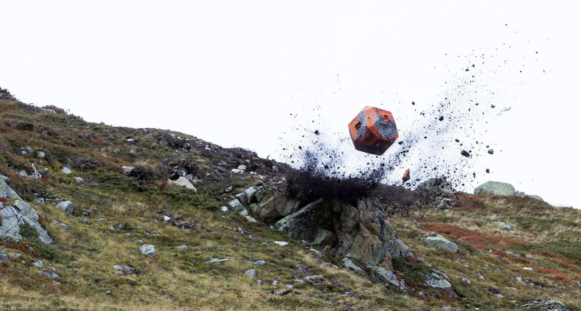 Ein großer, rot-schwarz lackierter Würfel stürzt auf einen felsigen Abhang und wirbelt vor dem klaren Himmel Erde und Trümmer in die Luft.