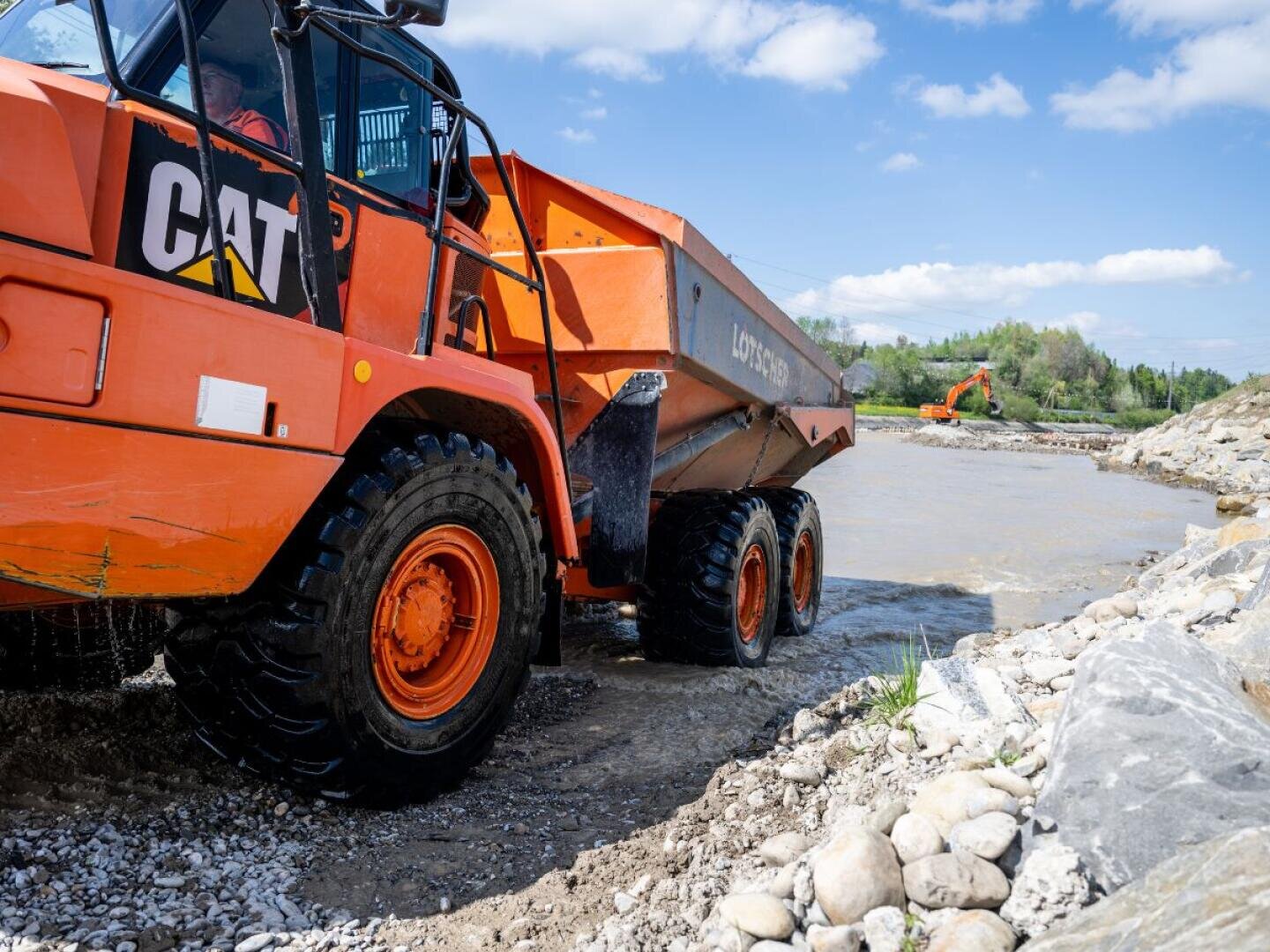 Ein orangefarbener Muldenkipper mit großen Reifen fährt durch flaches Wasser auf einer felsigen Baustelle, während im Hintergrund ein weiterer Bagger unter blauem Himmel mit vereinzelten Wolken zu sehen ist.